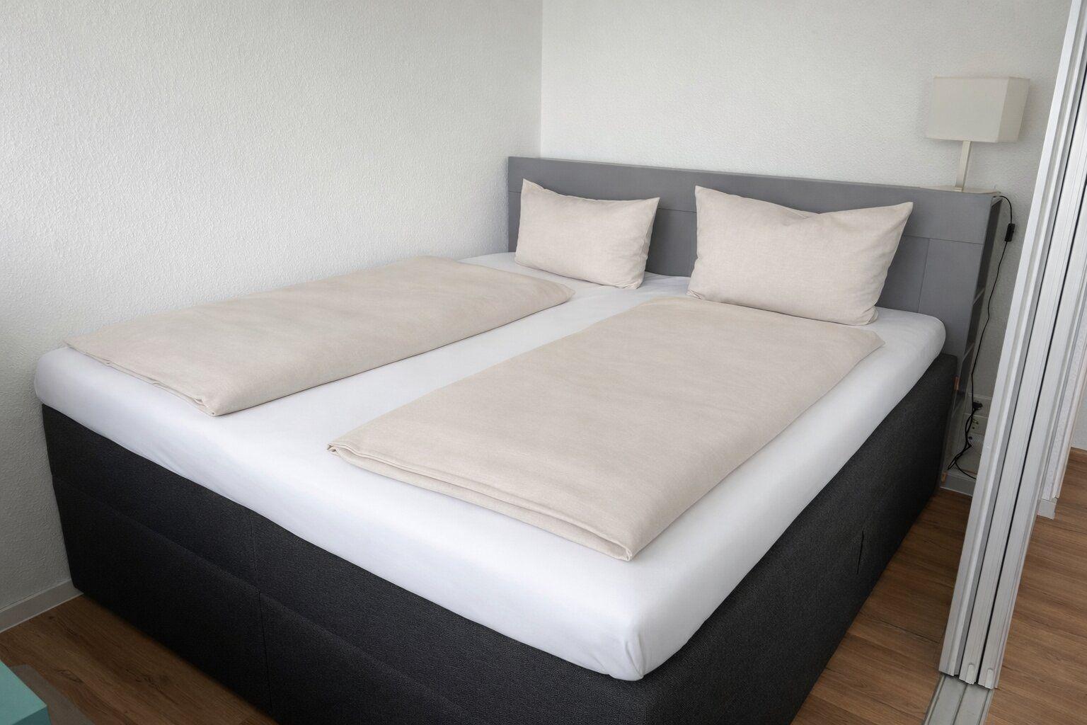 Doppelbett mit zwei Kissen und zwei Bettdecken, daneben Stehlampe und Holzboden.