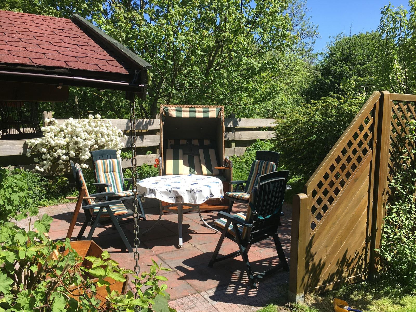 Terrasse mit Tisch, Stühlen und Liegestuhl unter Bäumen.