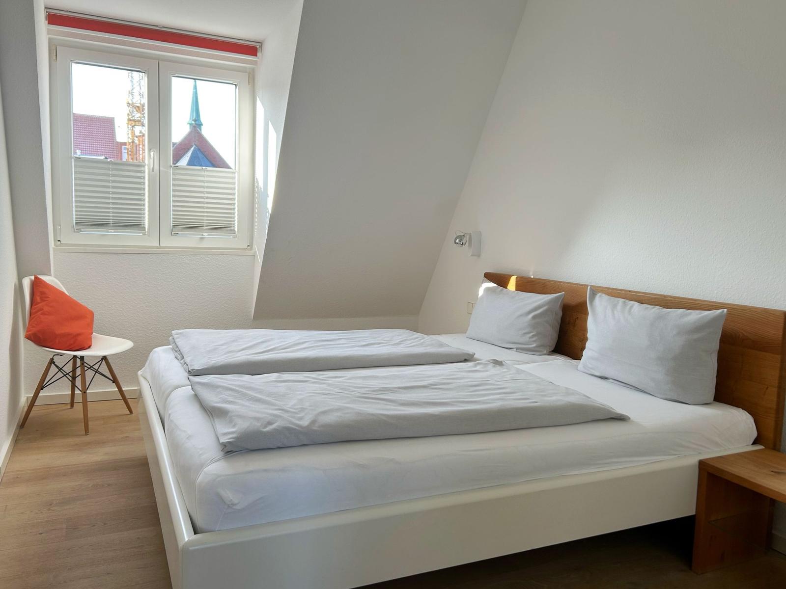 Schlafzimmer mit Doppelbett, Fenster und Stuhl mit Kissen.