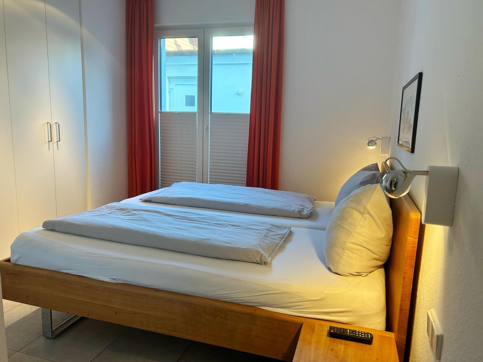 Schlafzimmer mit Doppelbett, Holzrahmen, Fenster mit roten Vorhängen und Wandlampe.