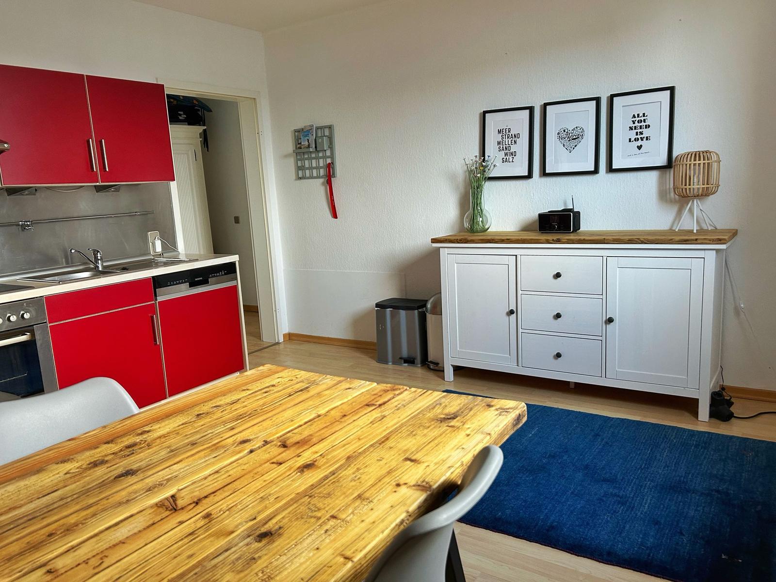 Küche mit roten Schränken und Holztisch. Weiße Kommode mit Bildern und Lampe.