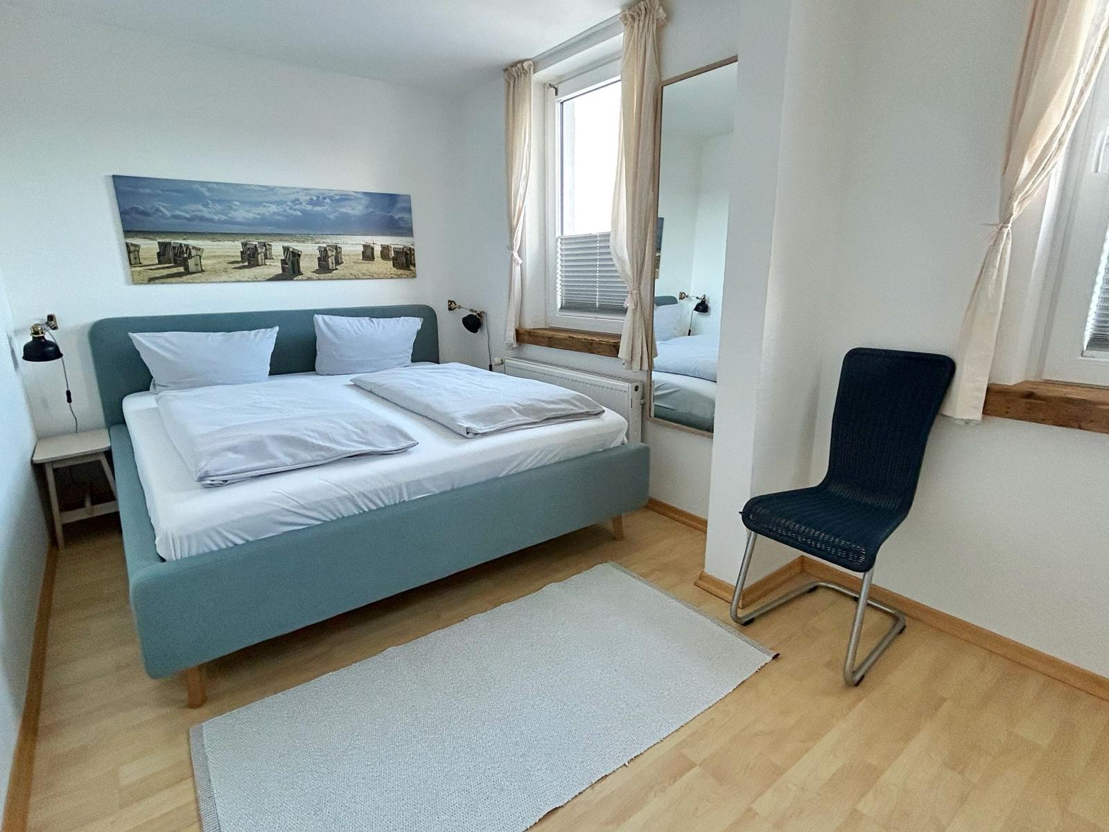 Schlafzimmer mit Bett, Stuhl und Fenster. Wandbild zeigt Strand.