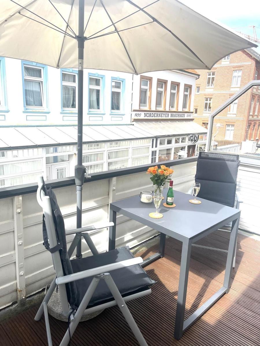 Terrasse mit Tisch, zwei Stühlen und Sonnenschirm. Blick auf Gebäude mit Schild.