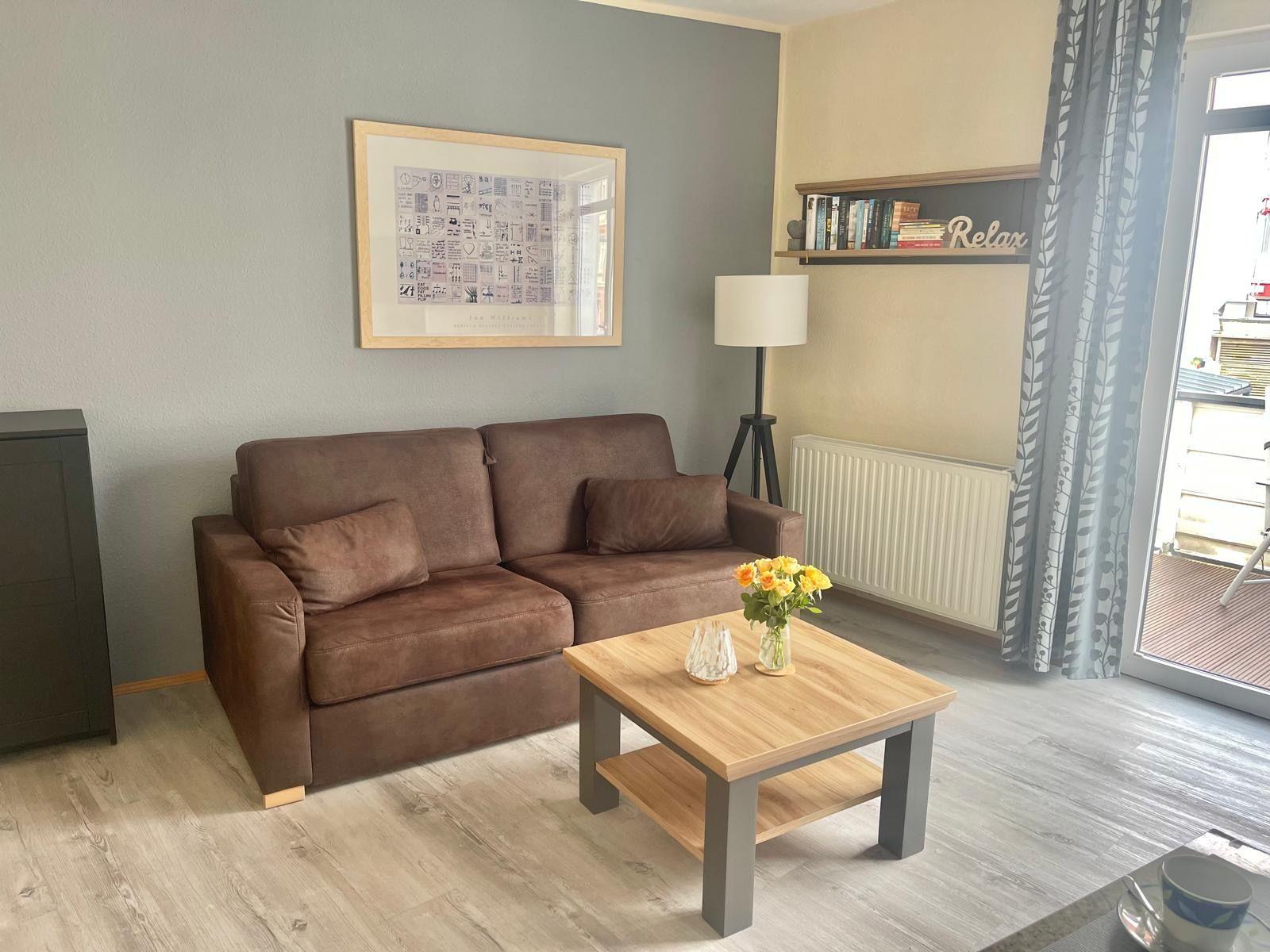 Ein Wohnzimmer mit braunem Sofa, Holztisch und Blumen. Hintergrund: Wandbild und Bücherregal.