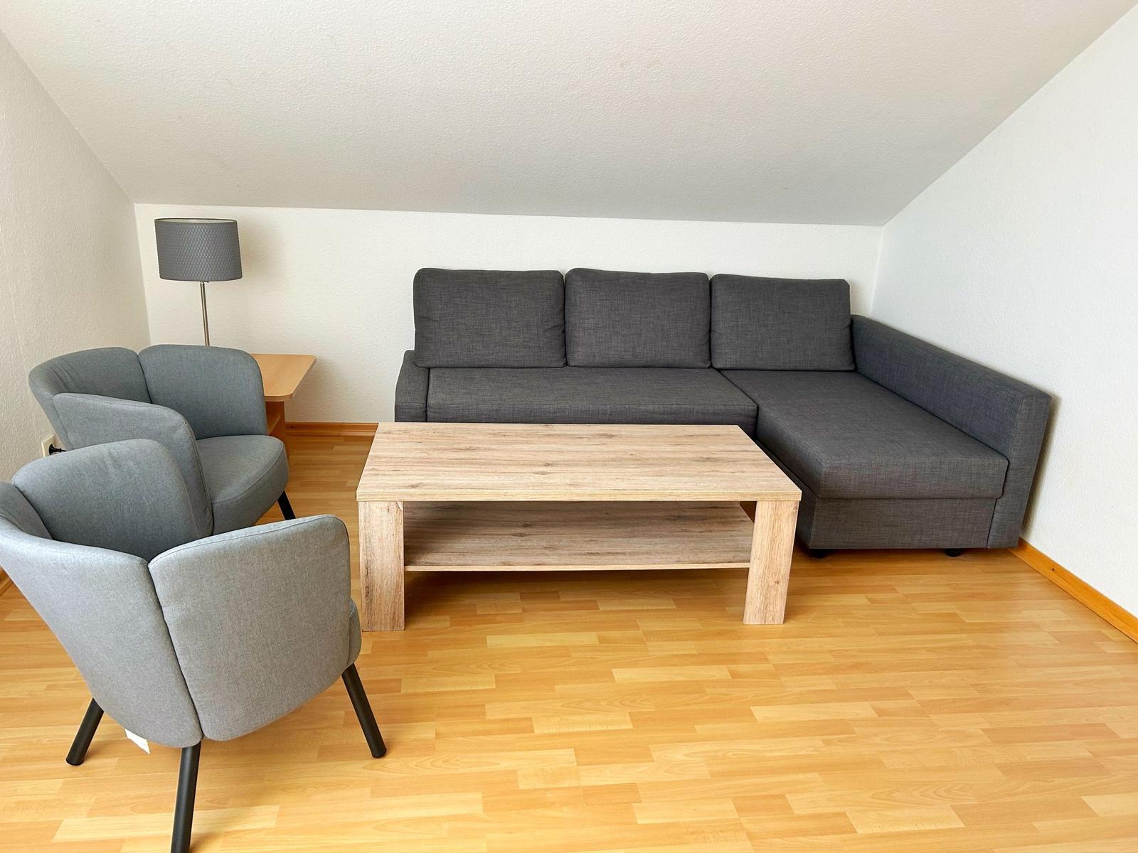 Wohnzimmer mit grauem Sofa, zwei Sesseln und Holztisch auf Parkettboden.