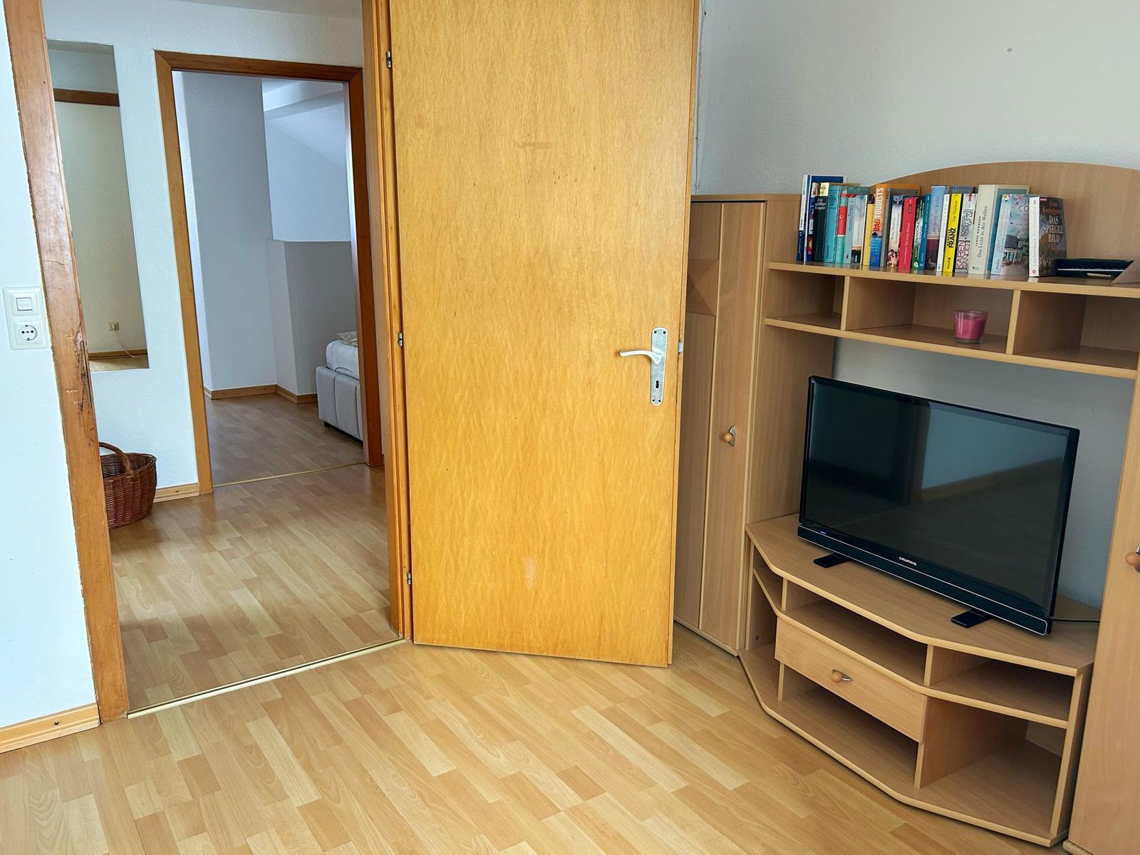 Wohnzimmer mit Fernseher und Bücherregal. Durchgang zu Schlafzimmer mit Sofa.