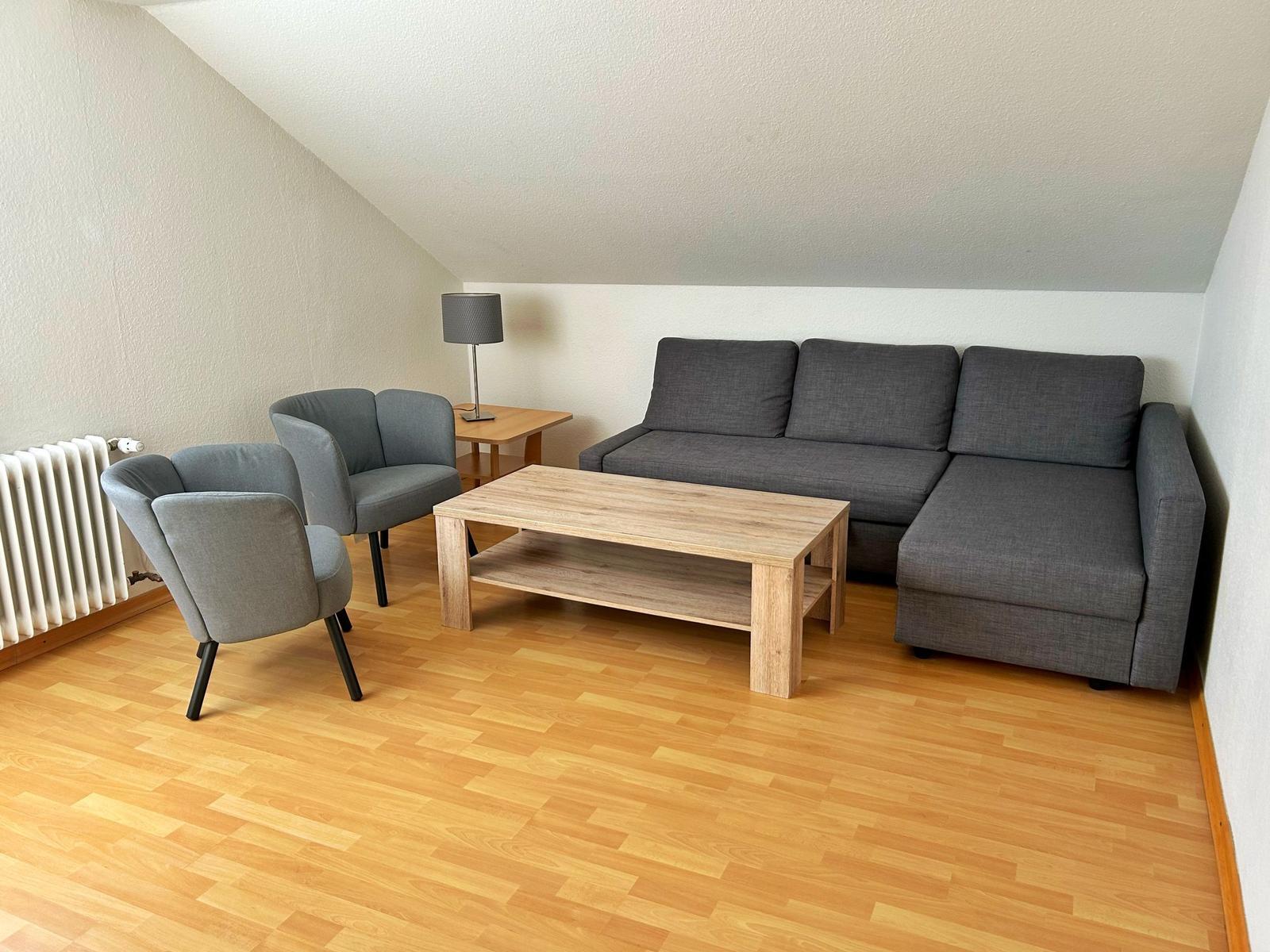 Wohnzimmer mit grauem Sofa, zwei Sesseln und Holztisch auf Parkettboden.