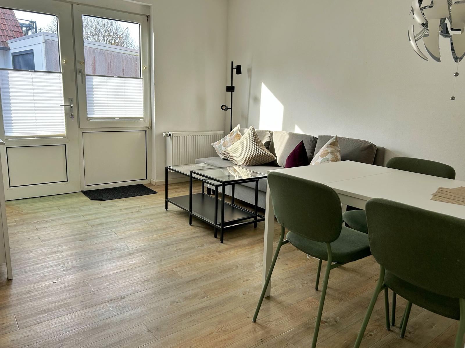 Wohn- und Essbereich mit Sofa, Tisch und Stühlen. Holzfußboden und Fenster mit Jalousien.