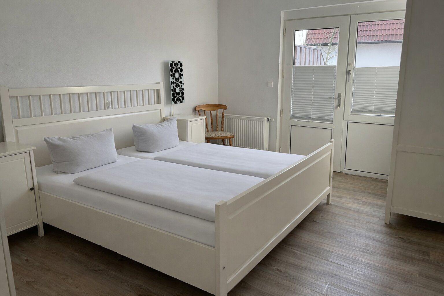 Zimmer mit Doppelbett, Nachttisch und Stuhl. Tür führt nach draußen.