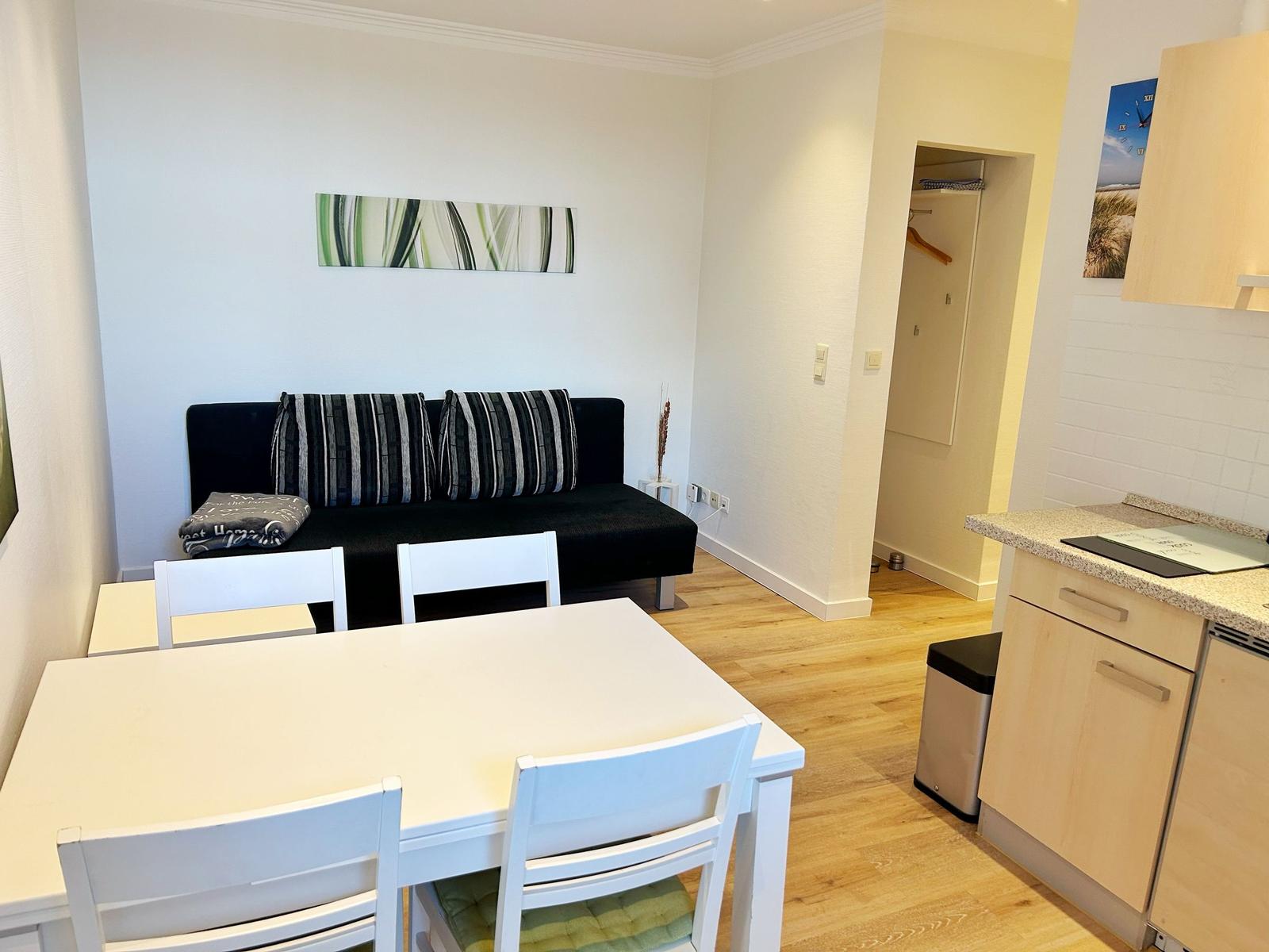 Wohnbereich mit Sofa, Esstisch und Küchenzeile. Holzboden und helle Wände.