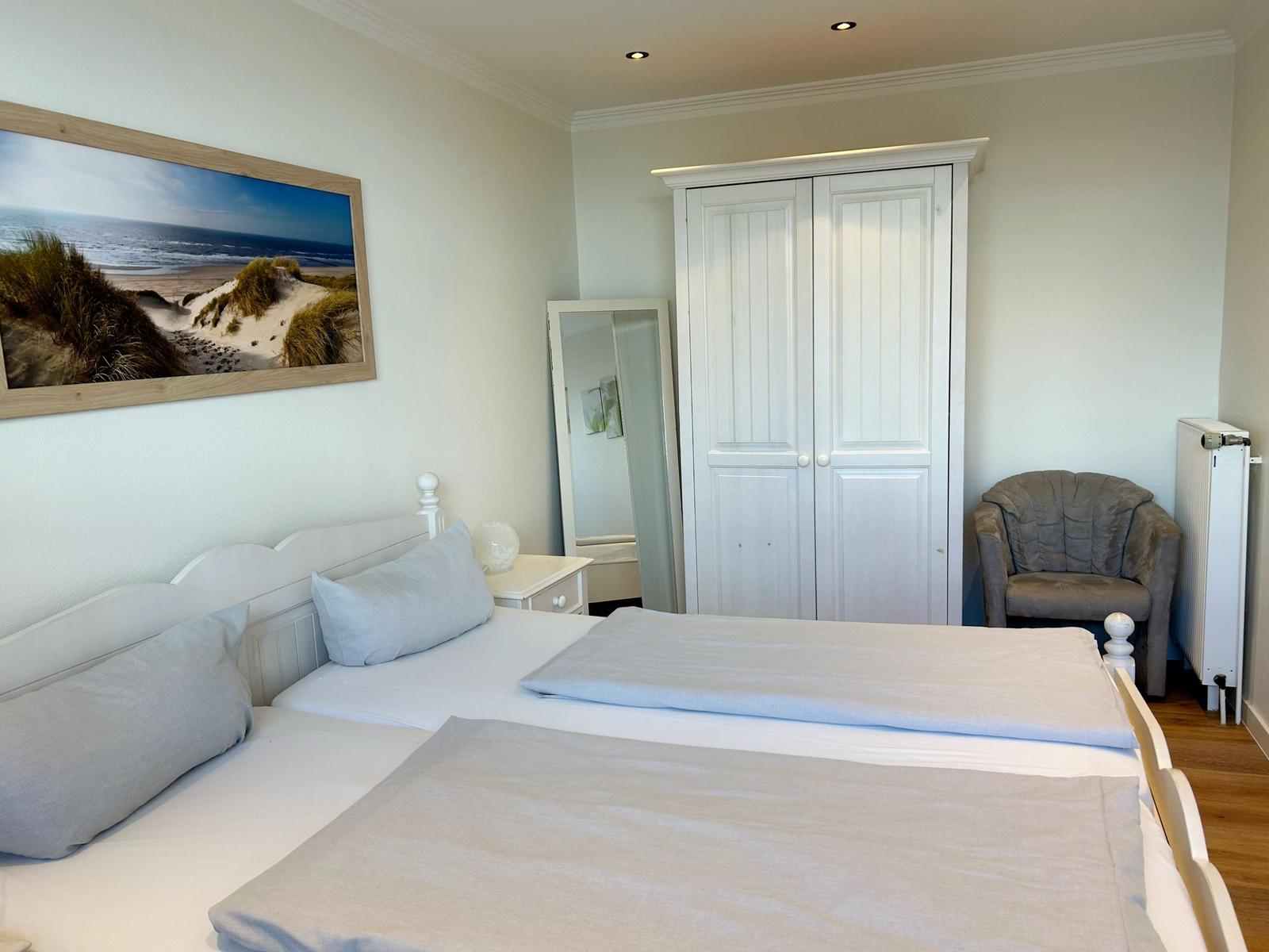 Schlafzimmer mit Doppelbett, Kleiderschrank, Sessel und Wandbild mit Strandmotiv.