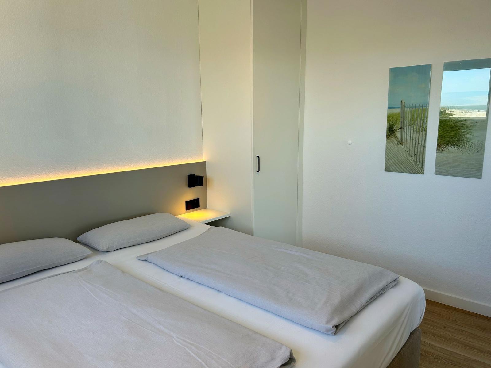Zimmer mit Doppelbett, Nachttisch und Wandbildern. Holzboden und Schrank.