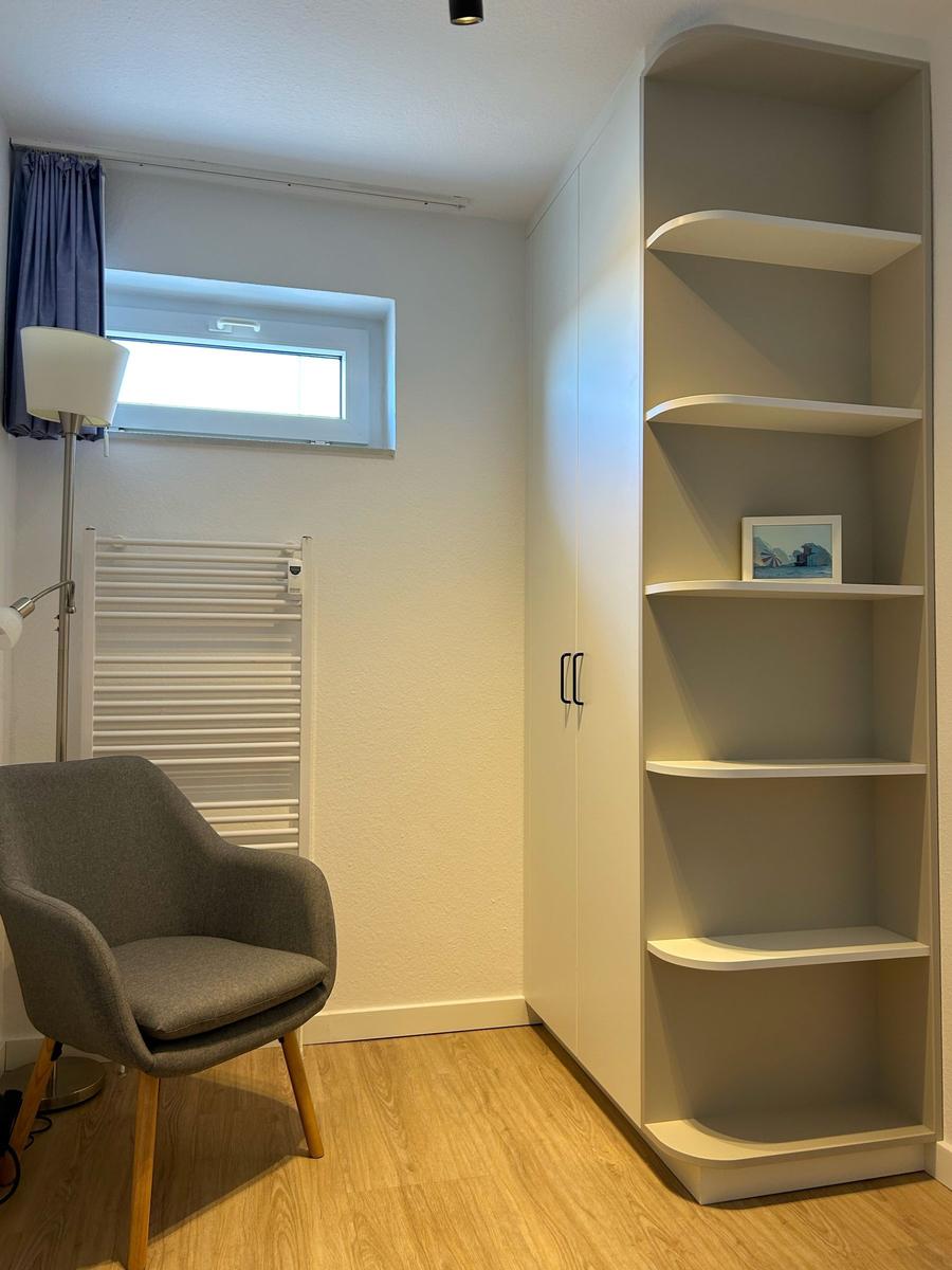 Ein Raum mit Stuhl, Bücherregal und Fenster mit Vorhang.