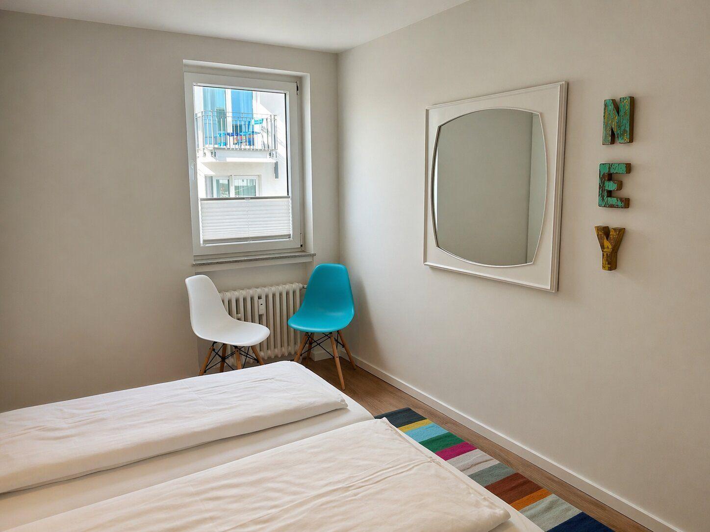 Zimmer mit Doppelbett, Fenster, Spiegel und bunten Stühlen.