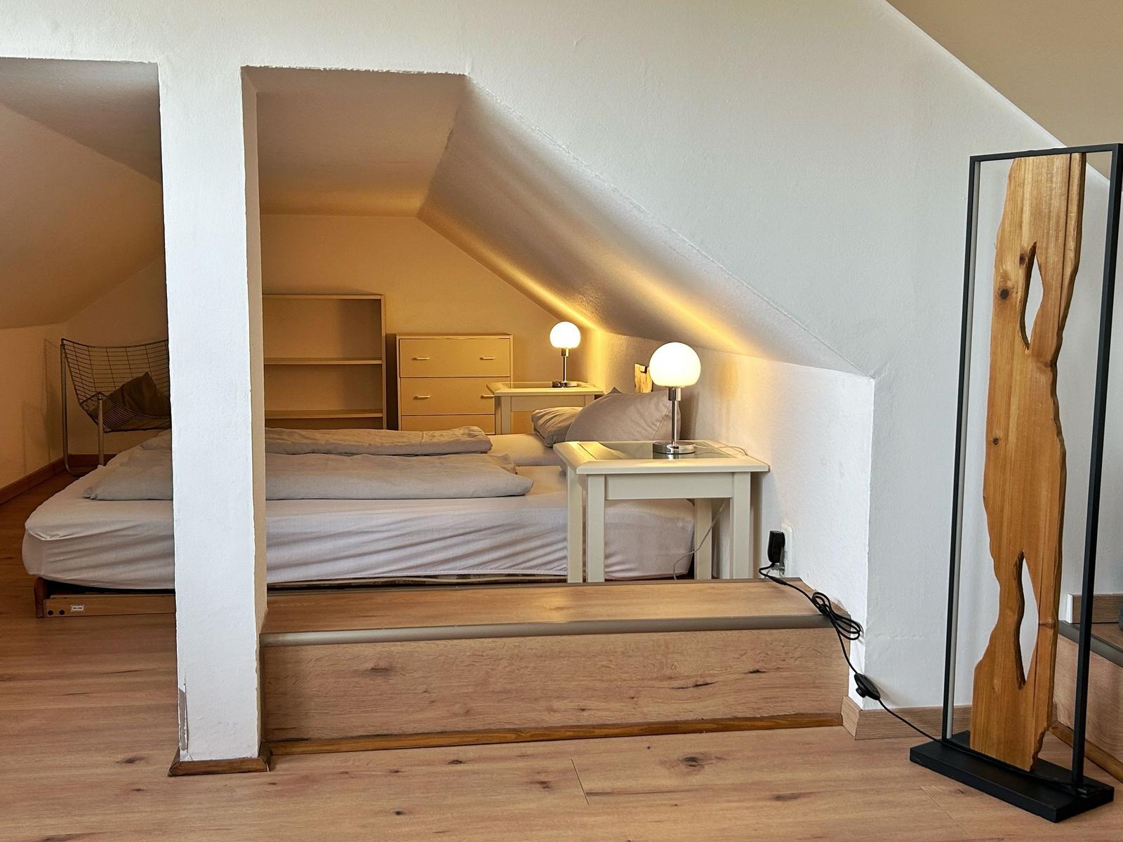 Schlafzimmer mit Dachschräge, Bett, Nachttisch und Lampen. Holzfußboden und Spiegel.
