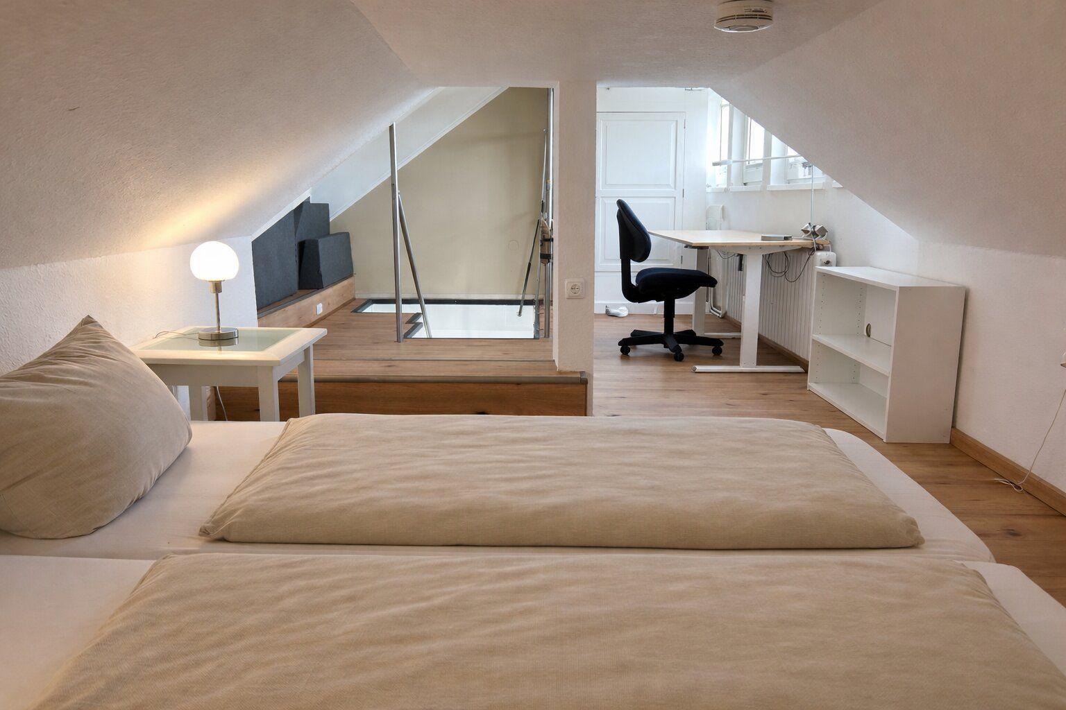 Schlafzimmer mit Bett, Nachttisch und Schreibtisch im Dachgeschoss. Blick auf die Treppe und den Flur.