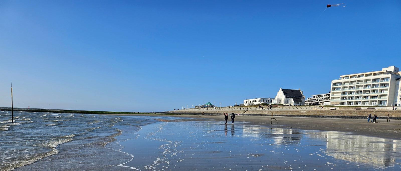 Strand mit Wellen, Menschen und Hotels im Hintergrund unter blauem Himmel.