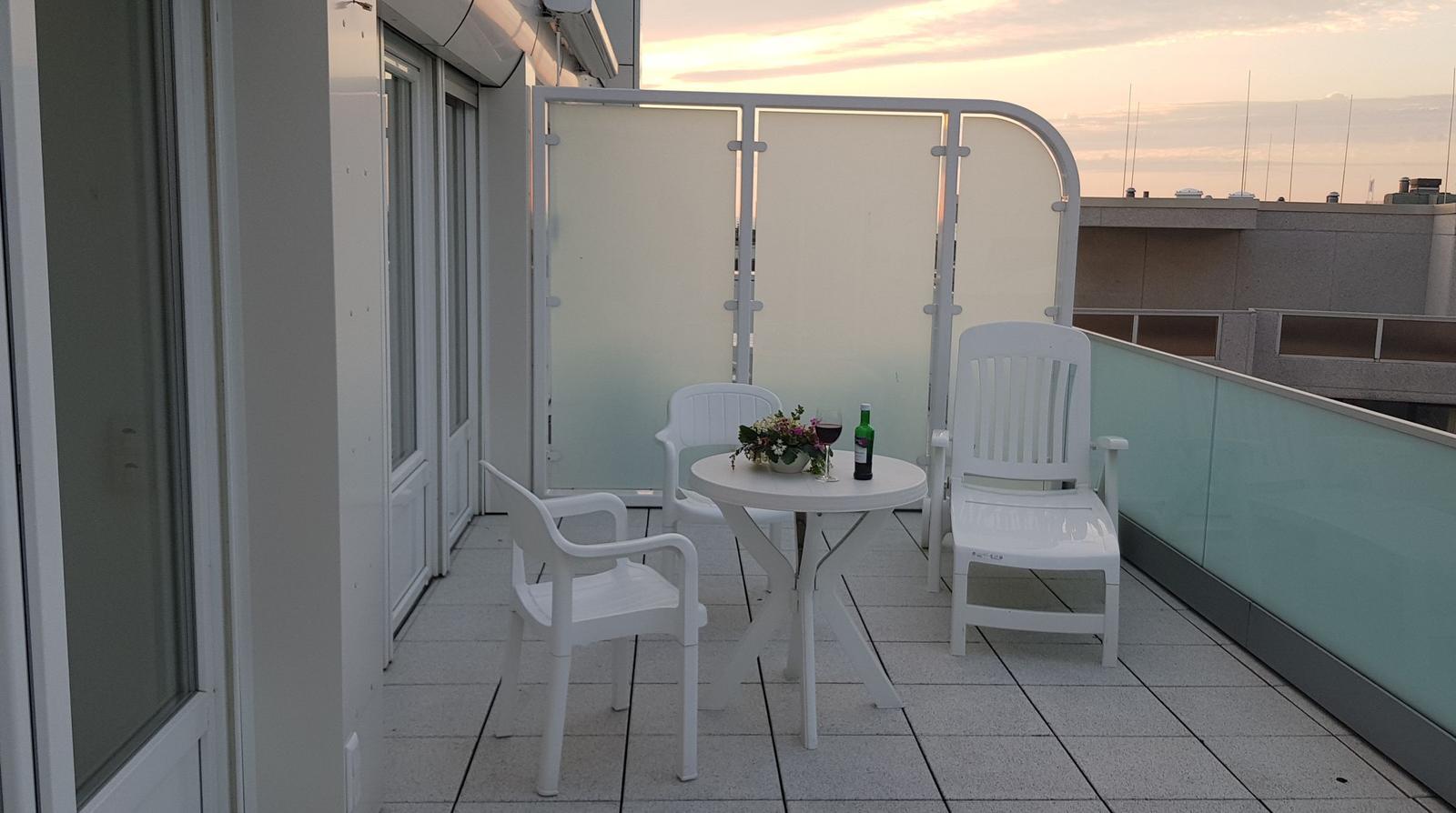 Terrasse mit weißem Tisch und Stühlen, Blumen und Weinflasche. Blick auf Gebäude und Himmel.