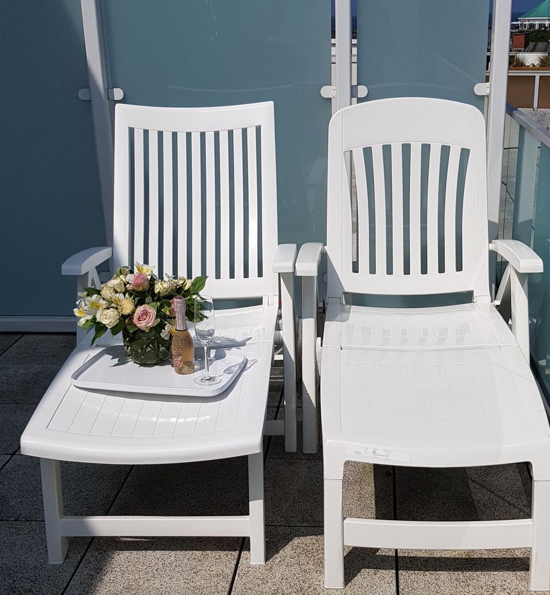 Zwei weiße Liegestühle auf Balkon mit Blumen und Flasche.