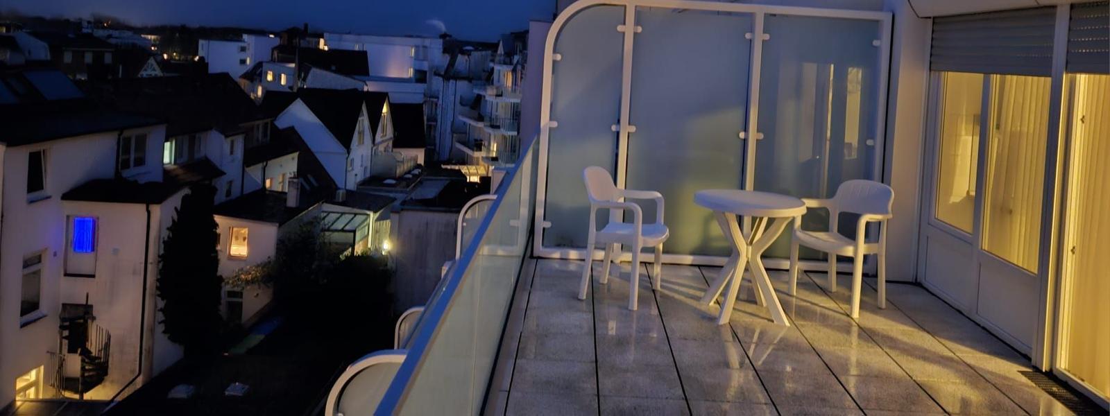 Terrasse mit Tisch und zwei Stühlen. Blick auf Wohngebiet bei Nacht.