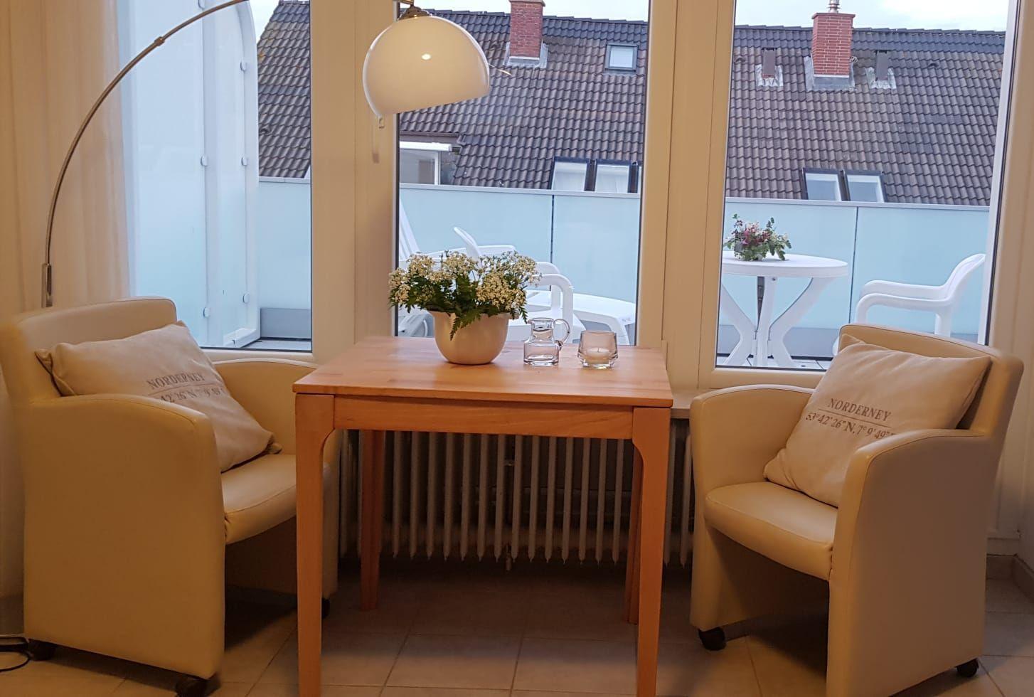 Zwei Sessel um einen Holztisch mit Blumen und Gläsern. Draußen ist eine Terrasse mit Tisch und Stühlen.