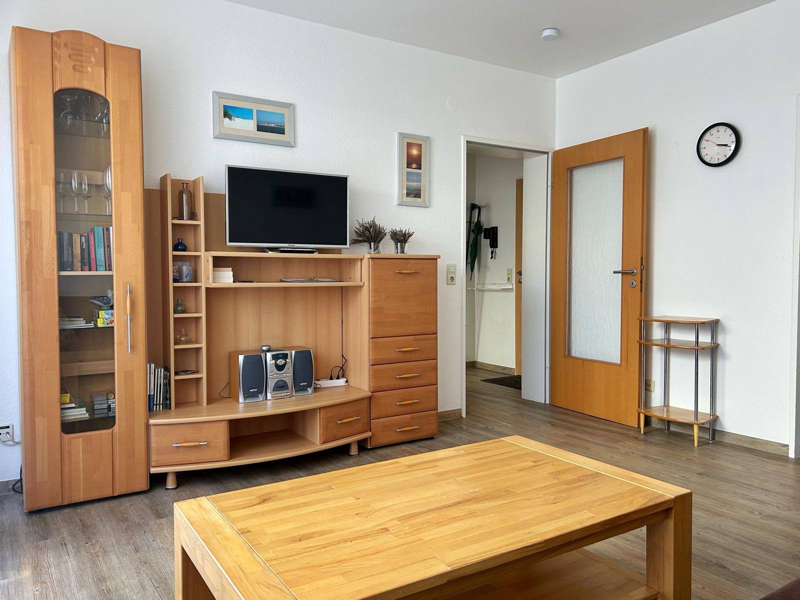 Wohnzimmer mit Holzmöbeln, Fernseher und Tisch. Tür führt in den Flur.