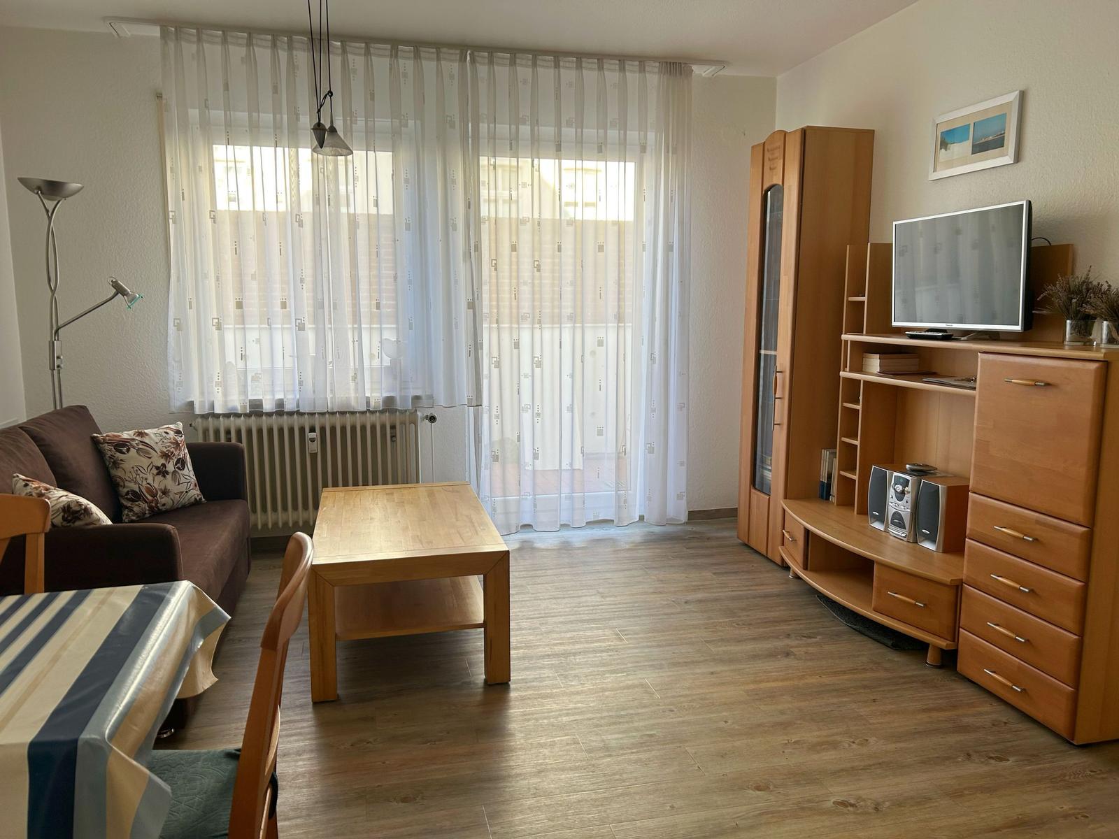 Wohnzimmer mit Sofa, Tisch, TV und Holzmöbel. Fenster mit weißen Vorhängen.