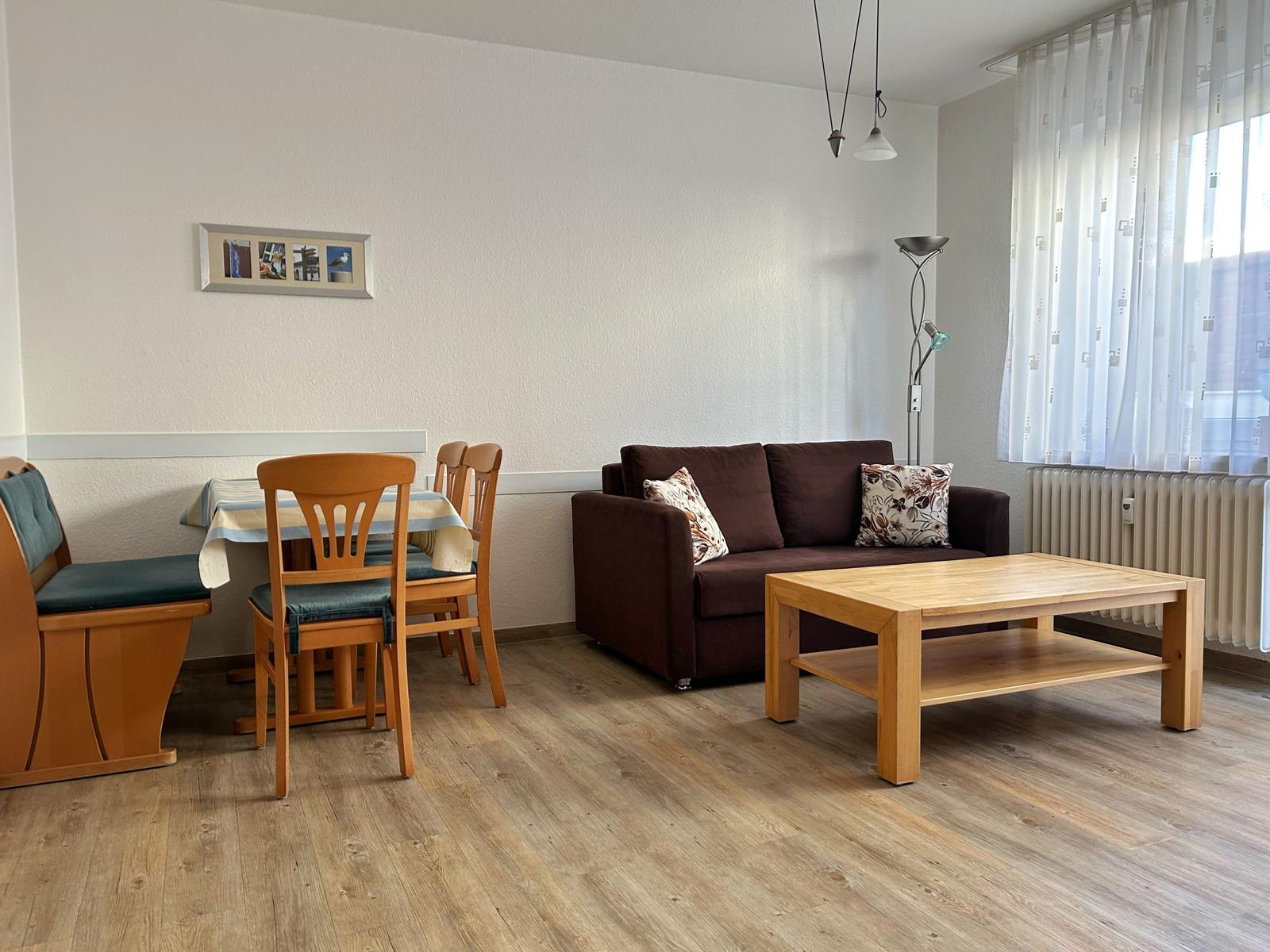 Wohn- und Essbereich mit Sofa, Tisch und Stühlen. Holzboden und Fenster mit Vorhängen.