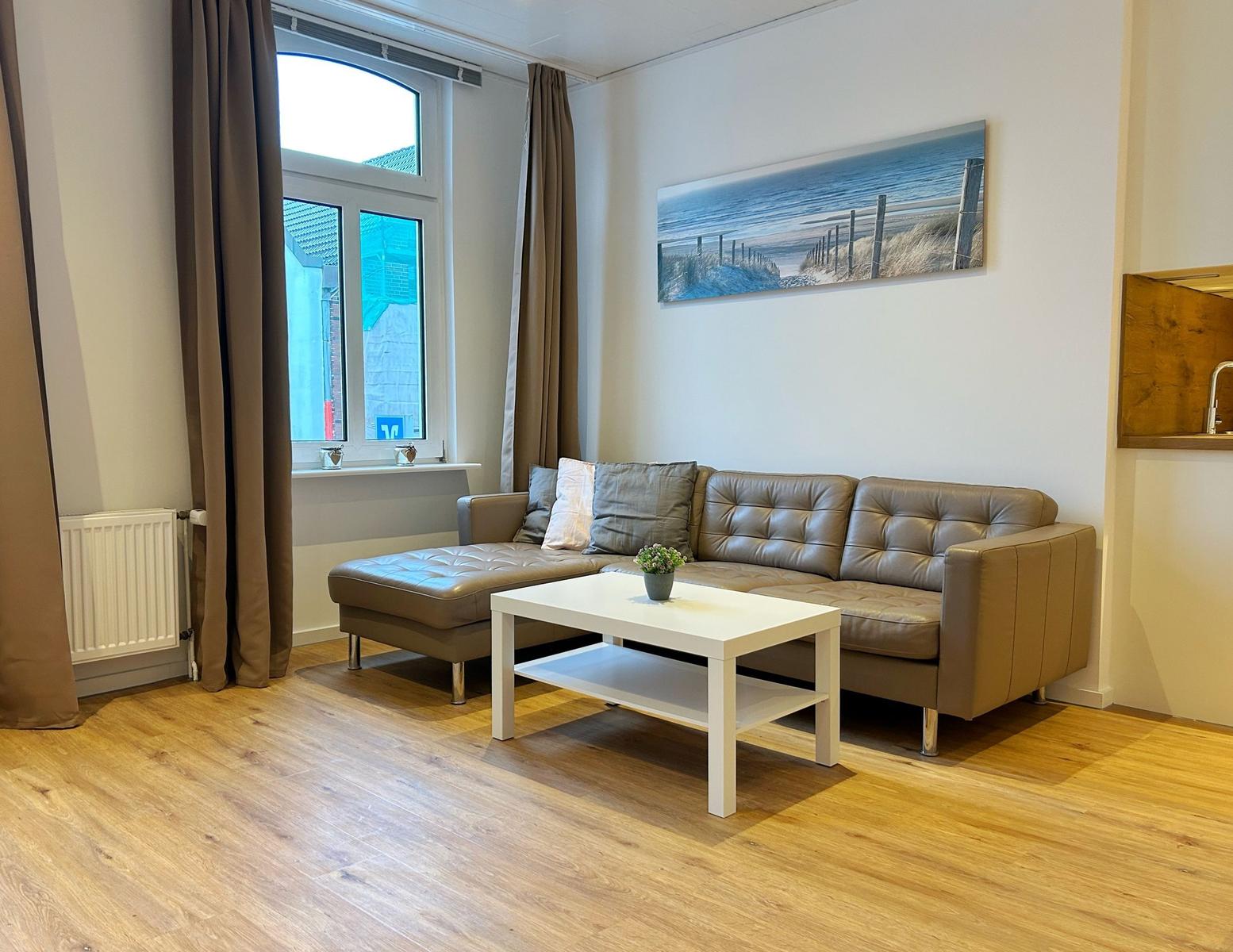Wohnzimmer mit braunem Sofa, weißem Tisch und Holzboden. Wandbild zeigt Strand.