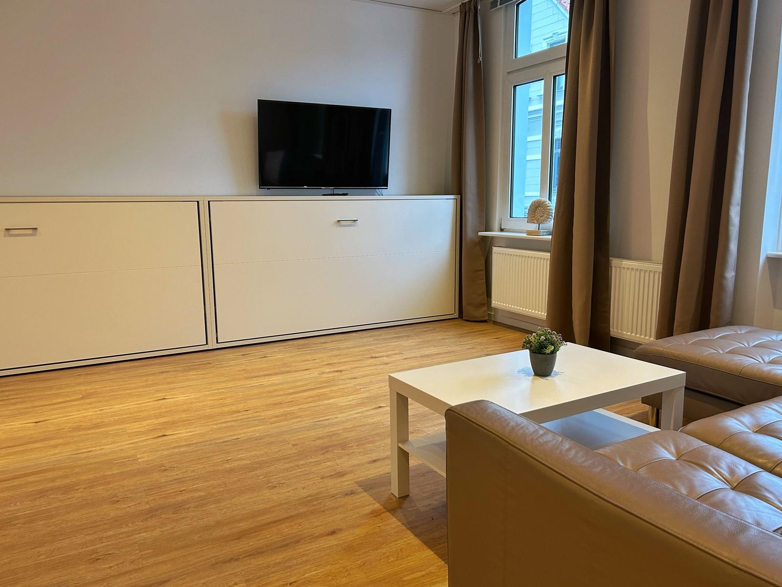 Wohnzimmer mit Holzboden, Sofa, TV und Fenster mit Vorhängen.