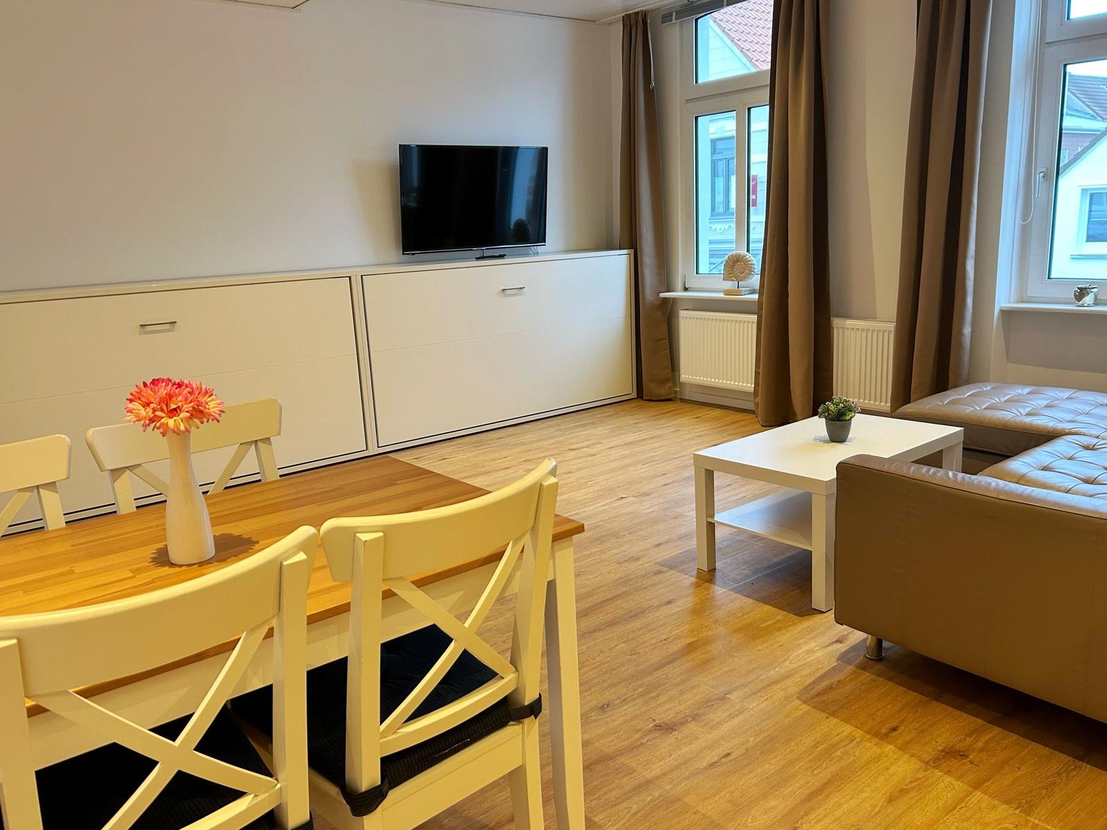 Wohn- und Essbereich mit Holzboden, TV, Sofa und Esstisch mit Stühlen.