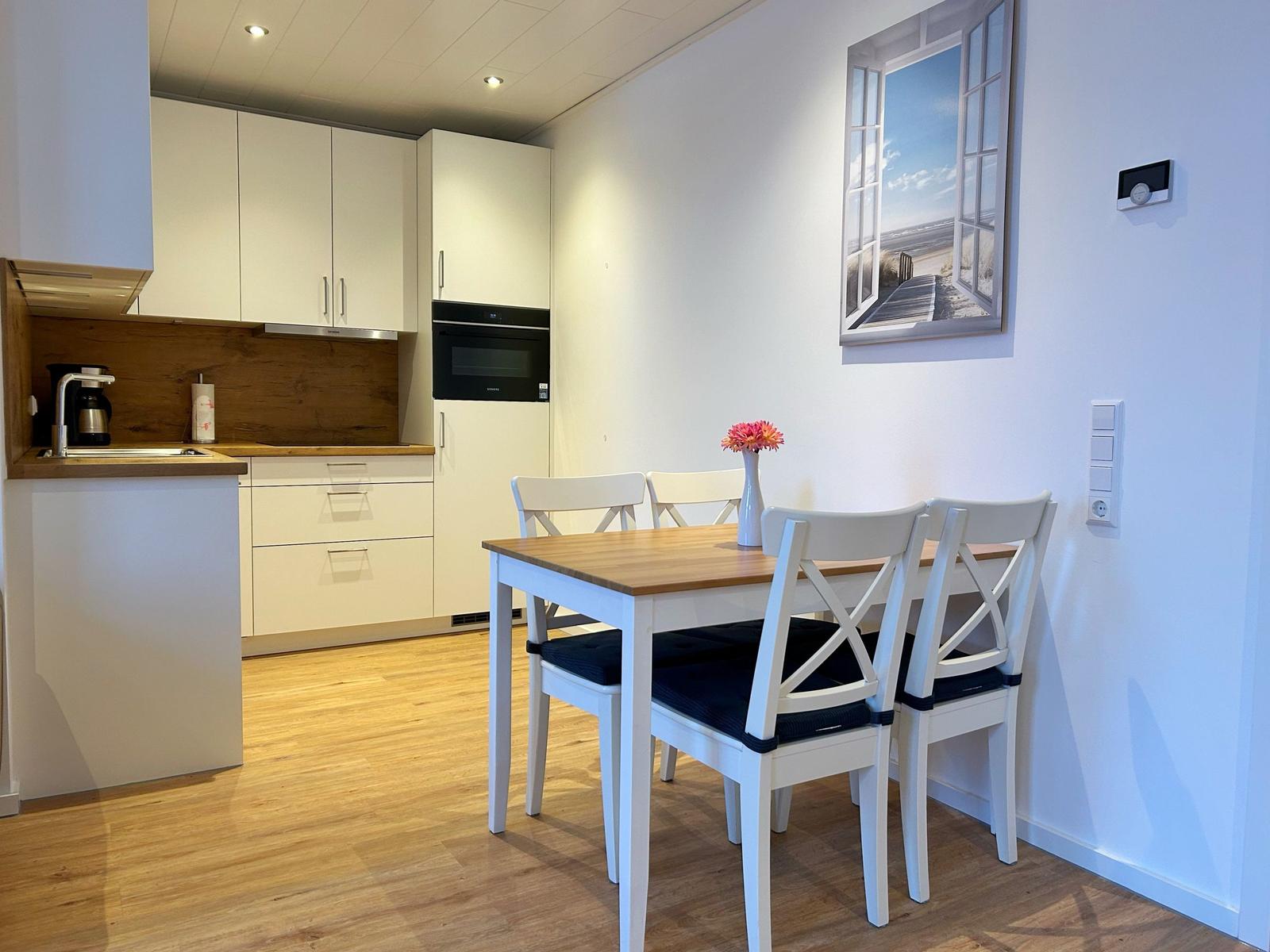 Küche mit Essbereich, weiße Möbel, Holzboden und Wandbild mit Strandmotiv.
