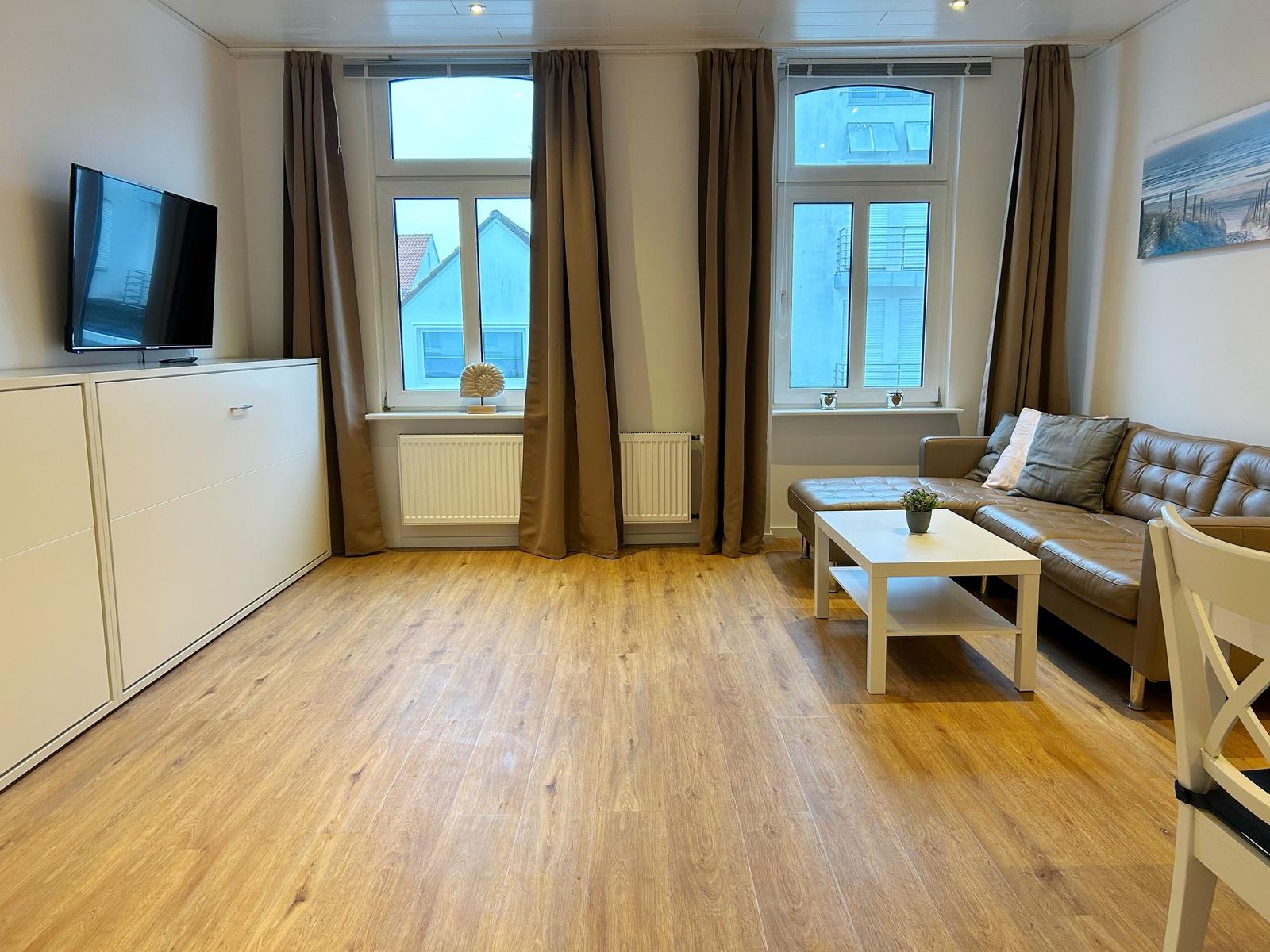 Wohnzimmer mit Ledersofa, TV und Holzboden. Fenster mit Vorhängen.