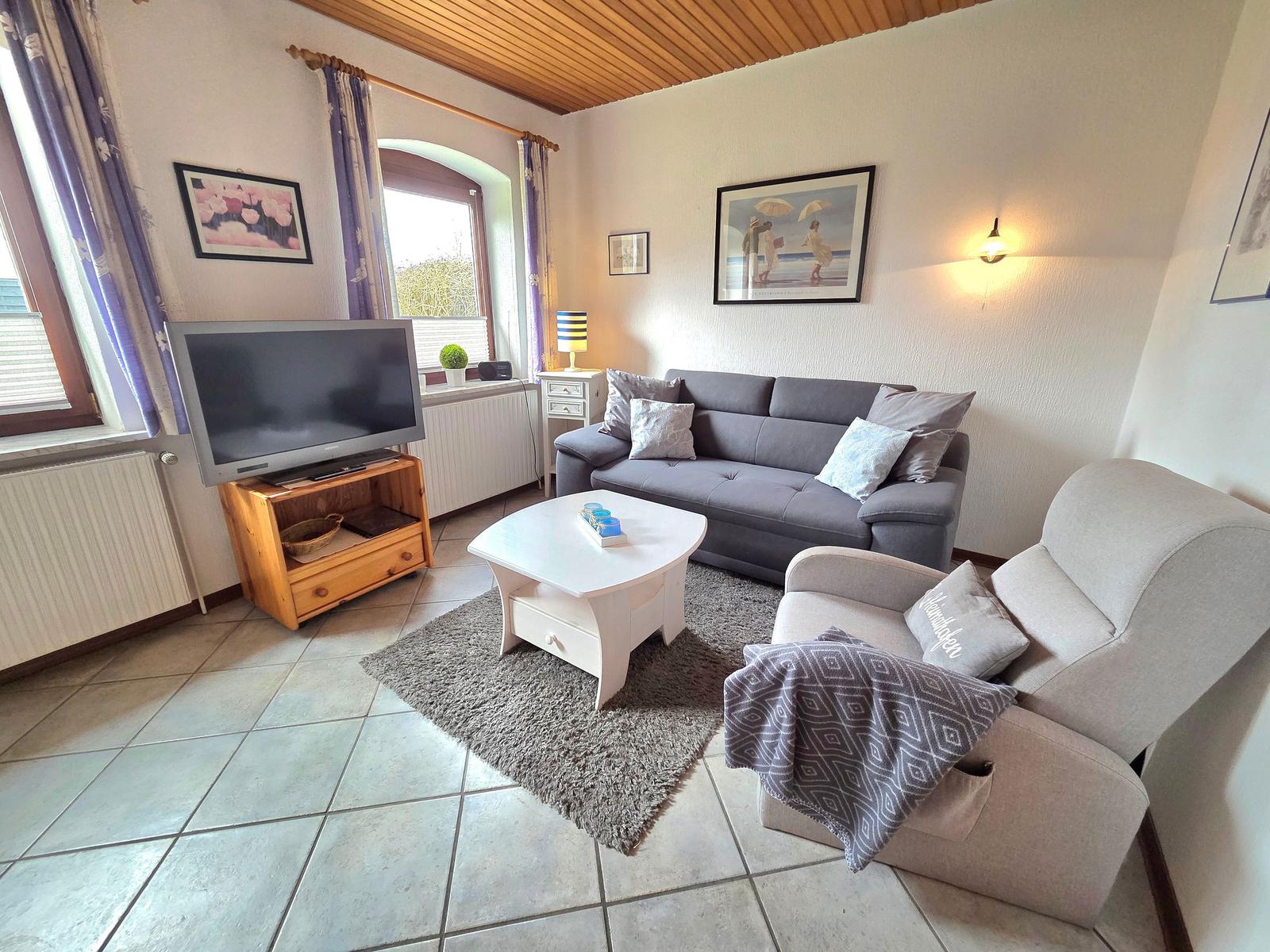 Wohnzimmer mit Sofa, Sessel, TV und Kaffeetisch. Holzdecke und Fenster mit Vorhängen.