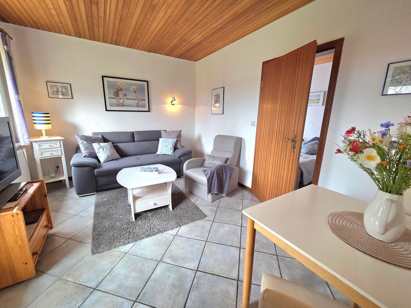 Wohnzimmer mit Sofa, Sessel und Esstisch. Tür führt ins Schlafzimmer.