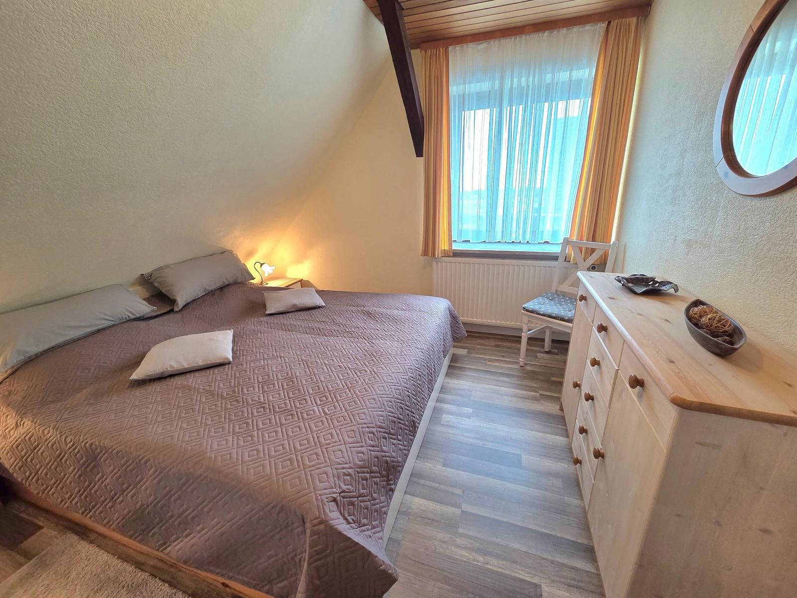 Schlafzimmer mit Doppelbett, Fenster, Schrank und Stuhl.