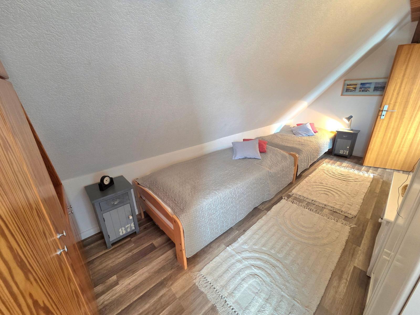 Zwei Einzelbetten mit grauer Bettwäsche und roten Kissen stehen in einem Dachzimmer mit Holzboden.