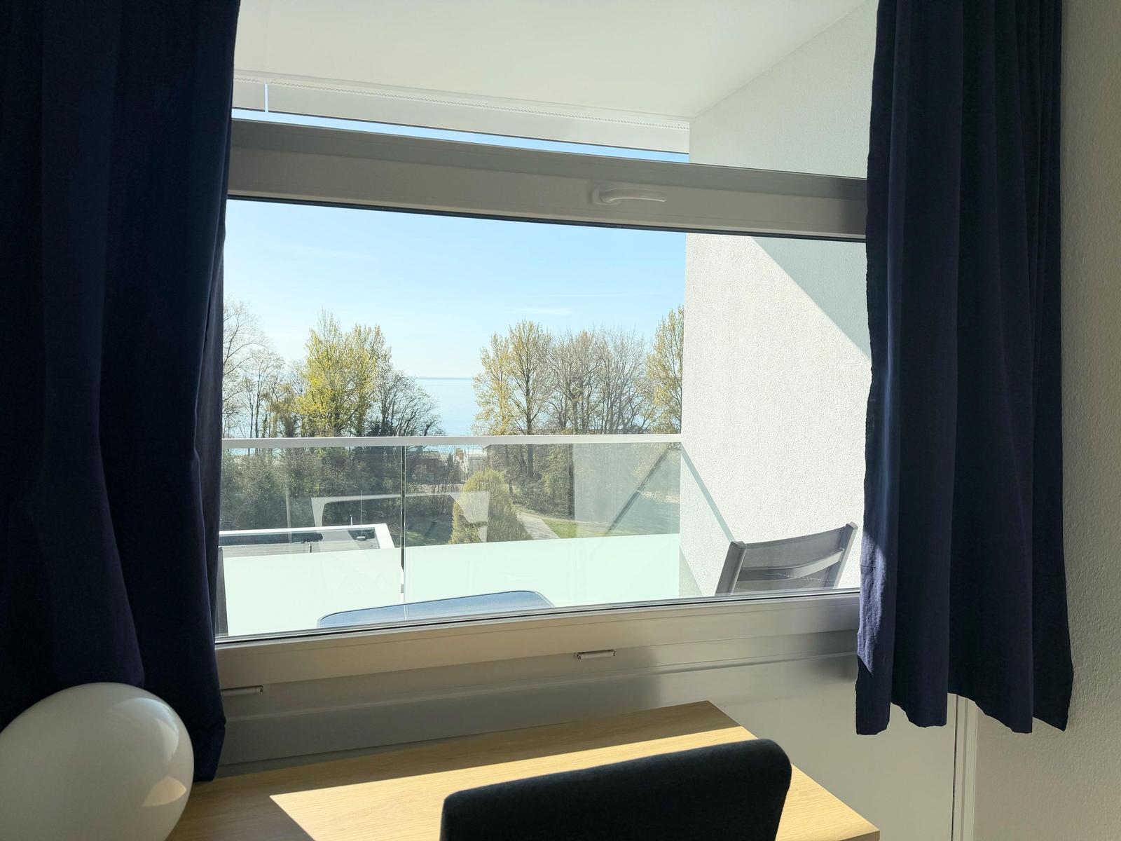 Blick aus dem Zimmer auf Balkon mit Glasgeländer und Baumreihe am Horizont.