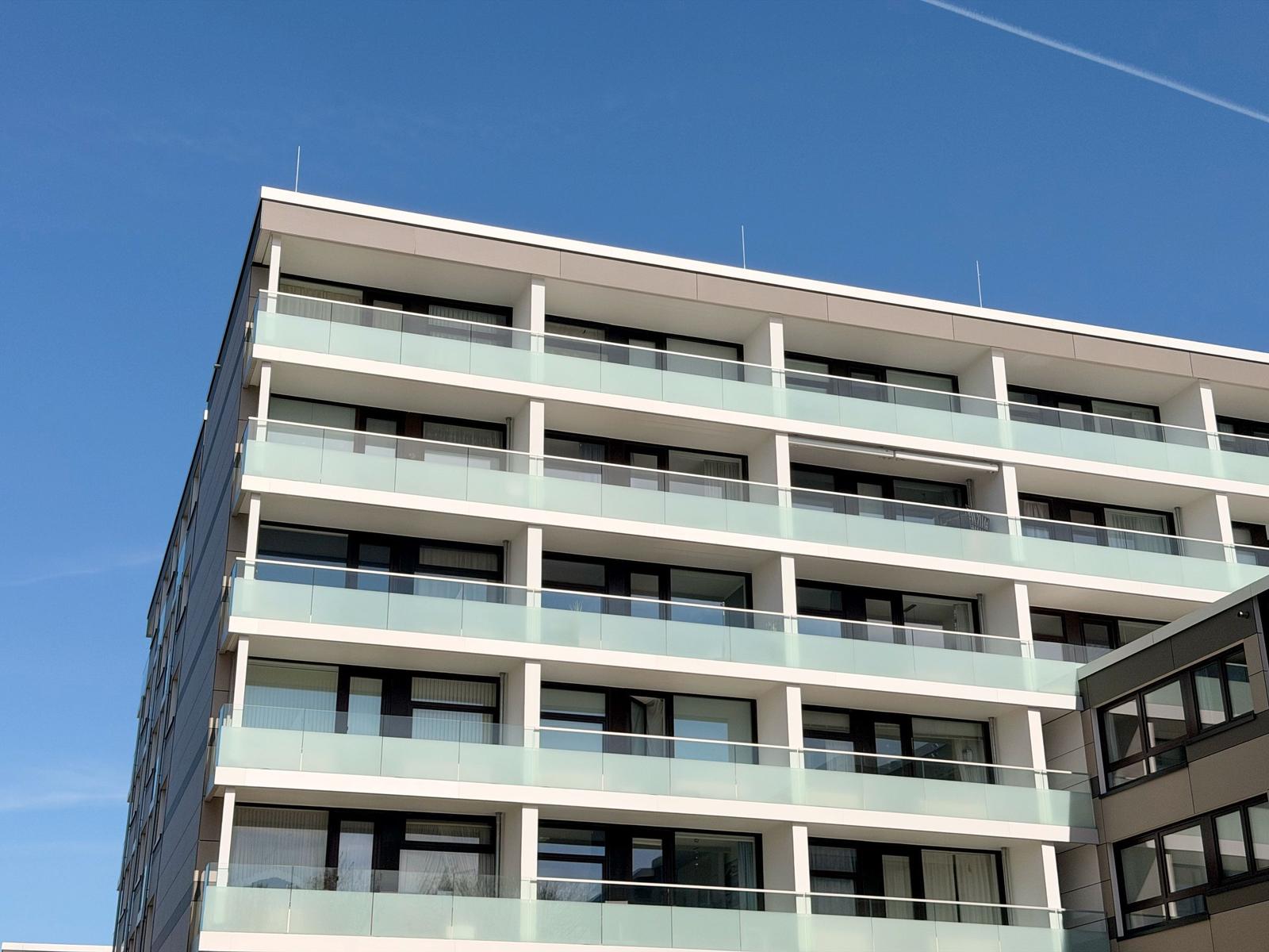Modernes Mehrfamilienhaus mit Glasgeländern und großen Fenstern unter blauem Himmel.
