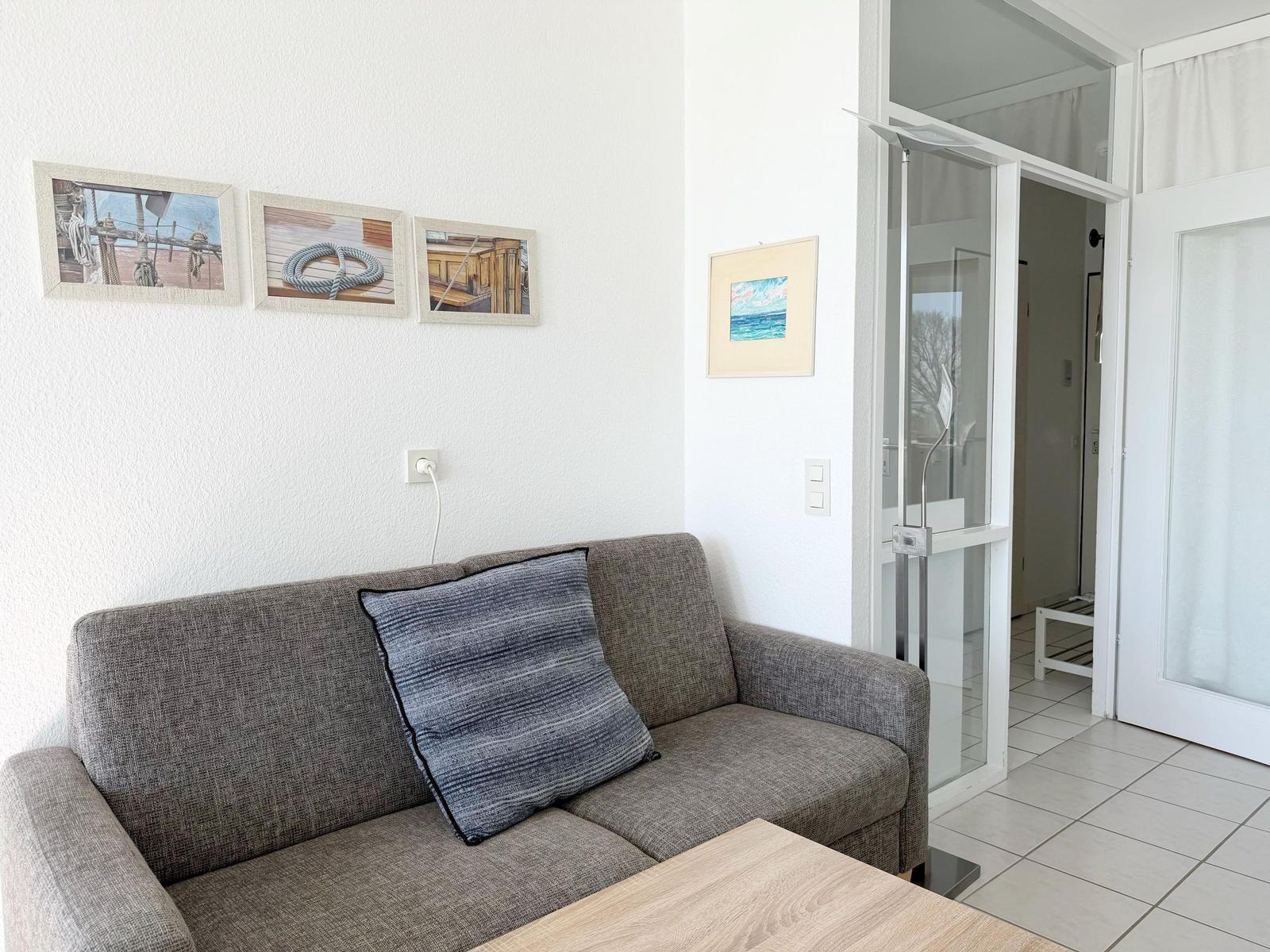 Ein Wohnbereich mit grauem Sofa, Kissen und Wandbildern. Durchgang zur Küche.
