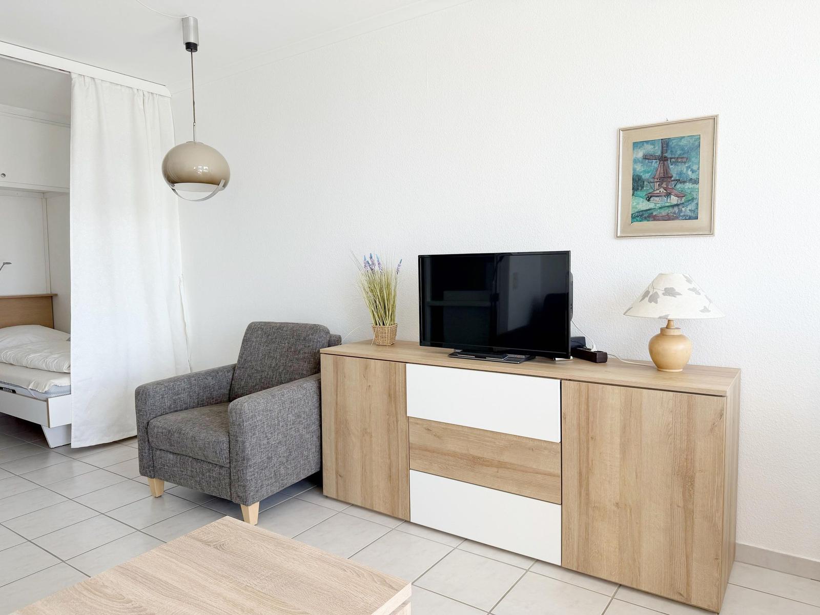 Wohnzimmer mit Sessel, TV-Schrank und Bett im Hintergrund.