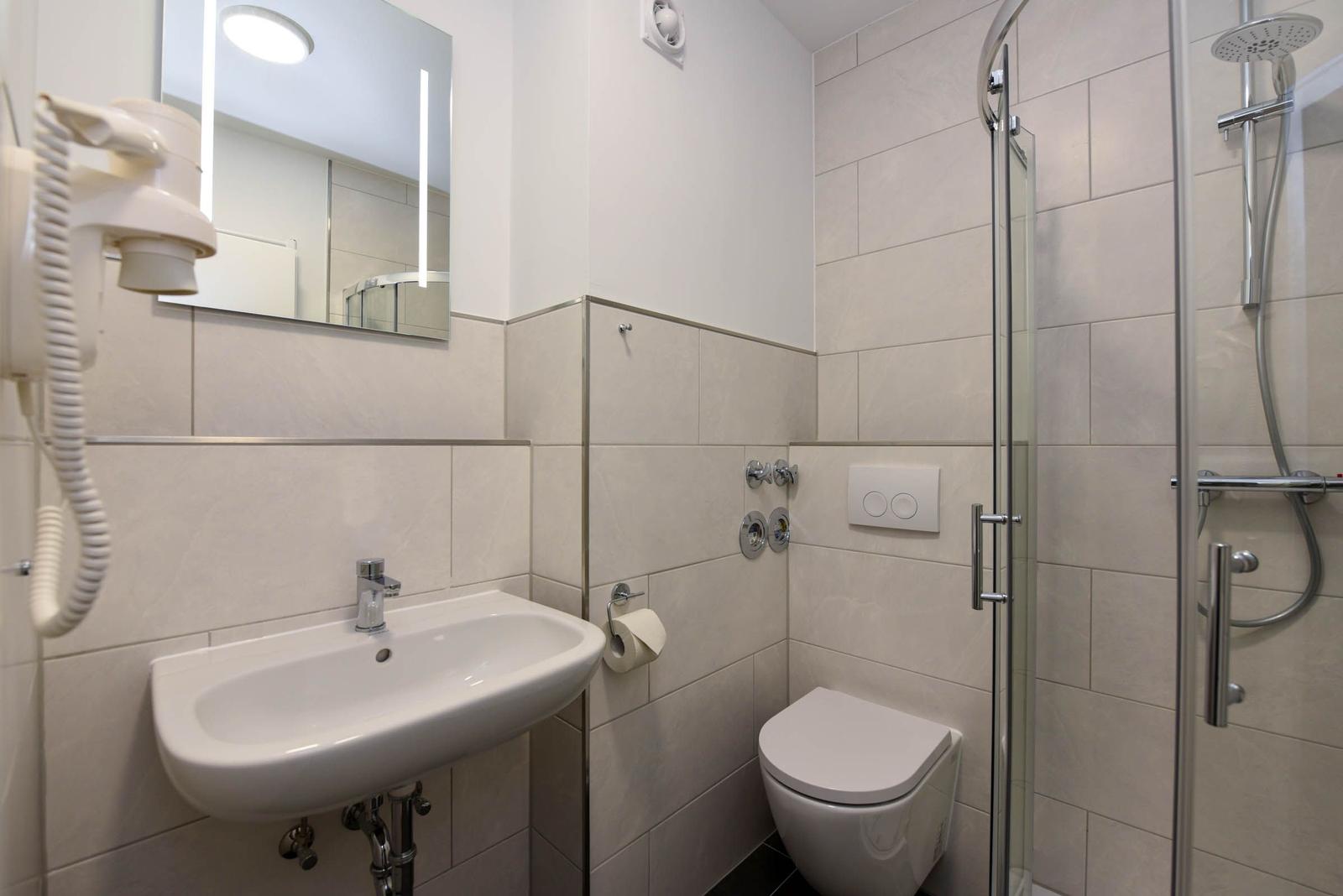 Badezimmer mit Dusche, Waschbecken und Toilette. Fliesenwand und Spiegel mit Beleuchtung.