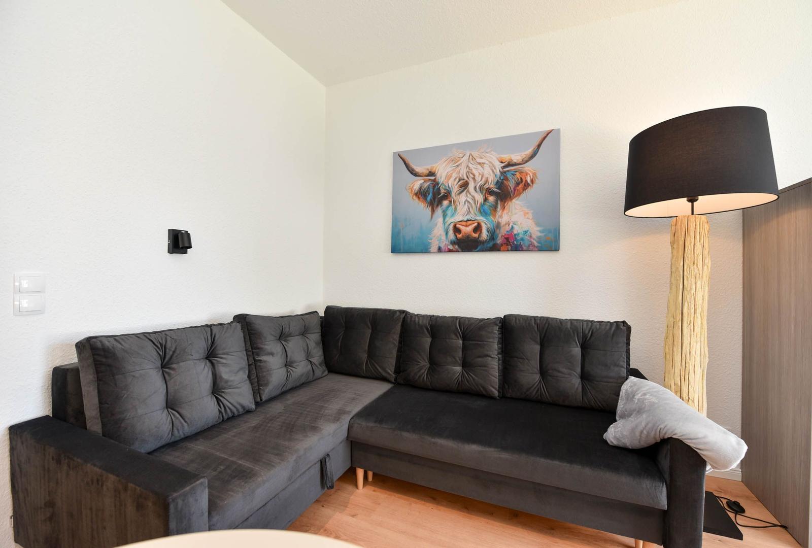 Ein Wohnzimmer mit grauem Ecksofa, Wandbild eines Hochlandrindes und Stehlampe.