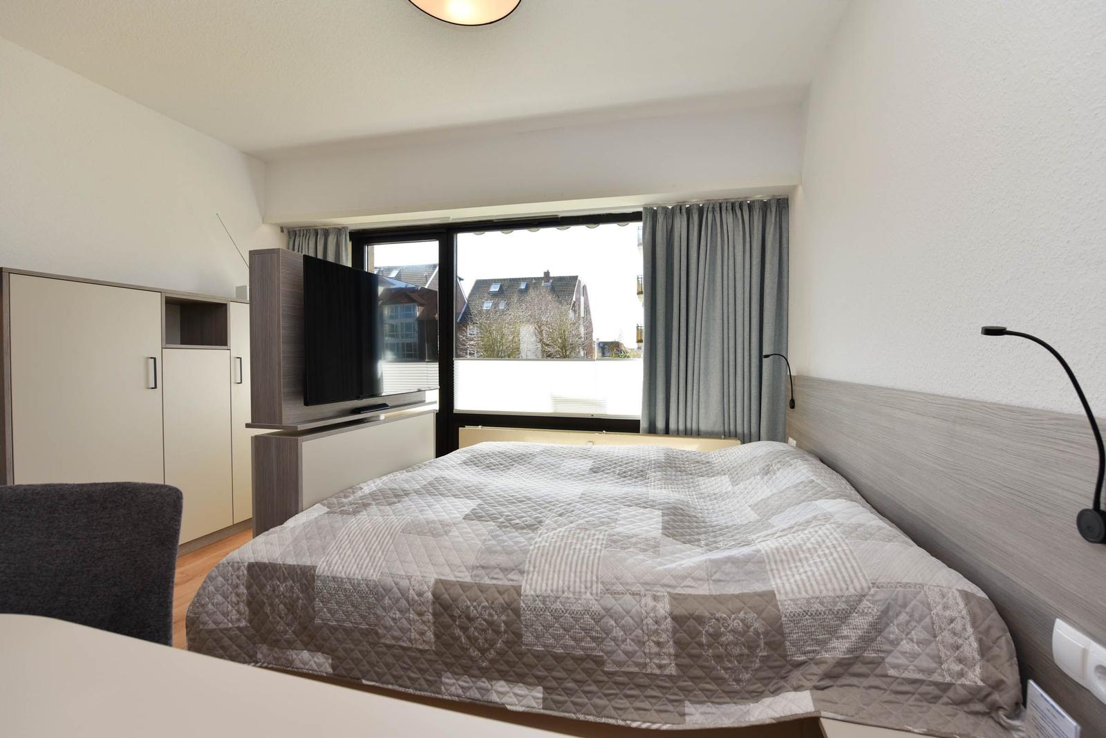 Schlafzimmer mit Bett, TV und Fenster mit Blick auf Gebäude.