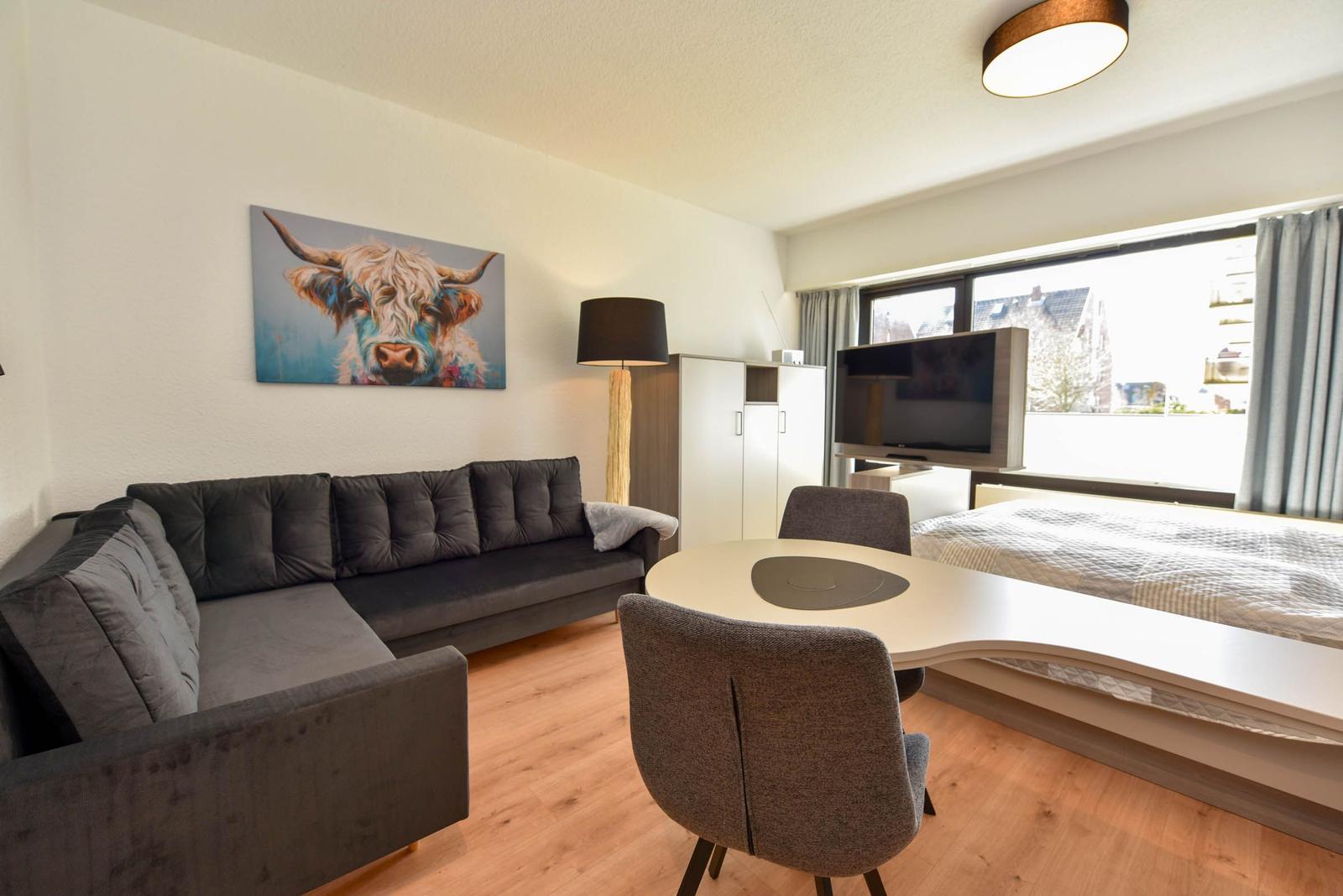 Wohnbereich mit Sofa, Esstisch und Bett. Wandbild eines Hochlandrindes.