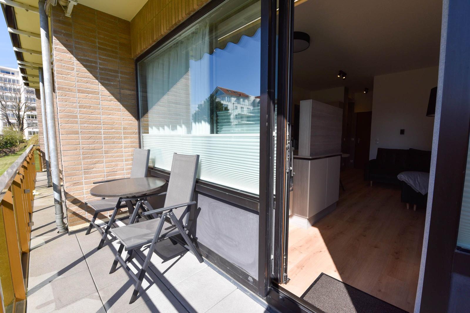 Terrasse mit Tisch und zwei Stühlen. Durch Glastür ins Innere mit Wohn- und Küchenbereich.