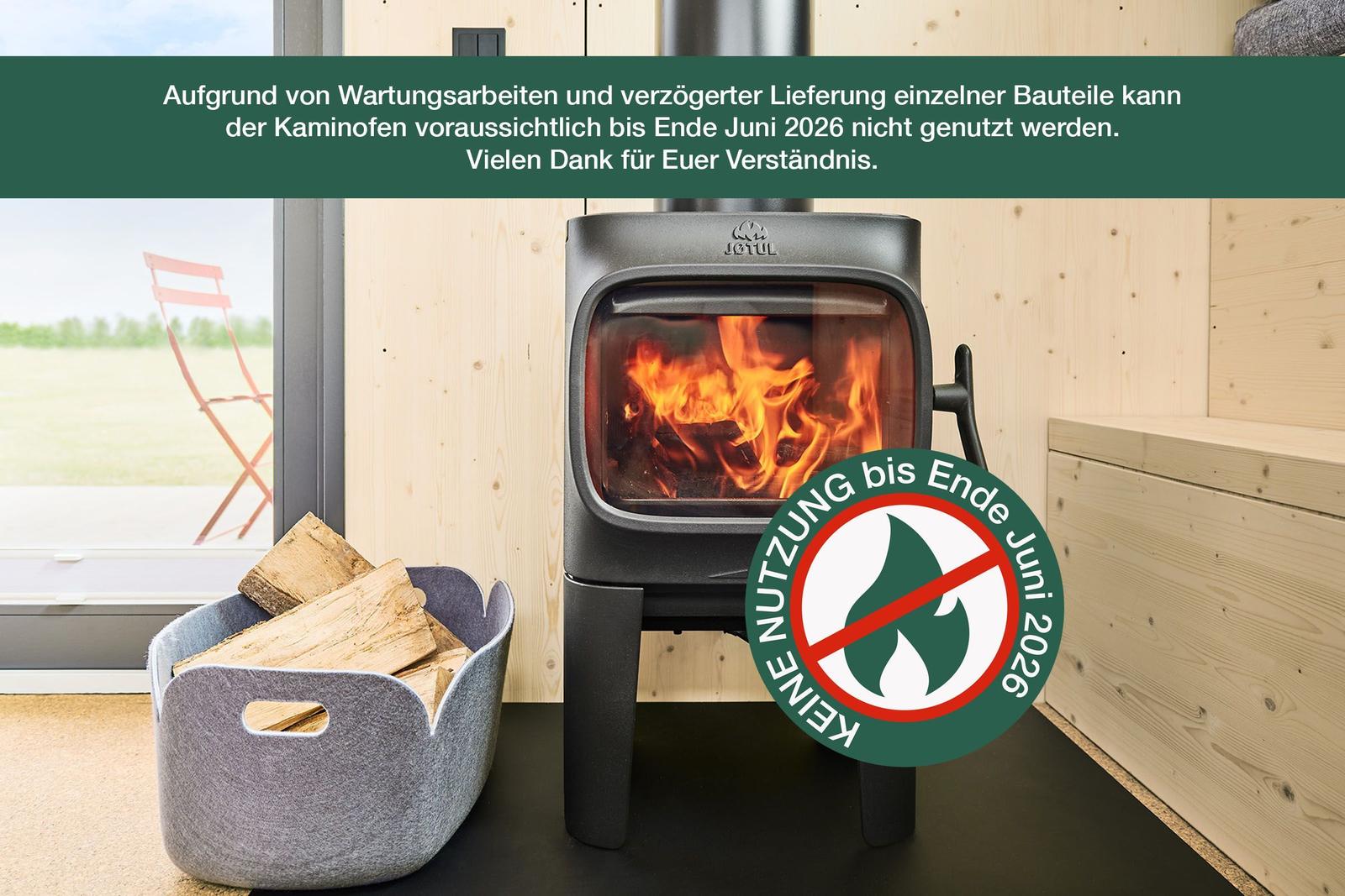 Kaminofen mit brennendem Holz, daneben Korb mit Brennholz und Fensterblick auf Garten.