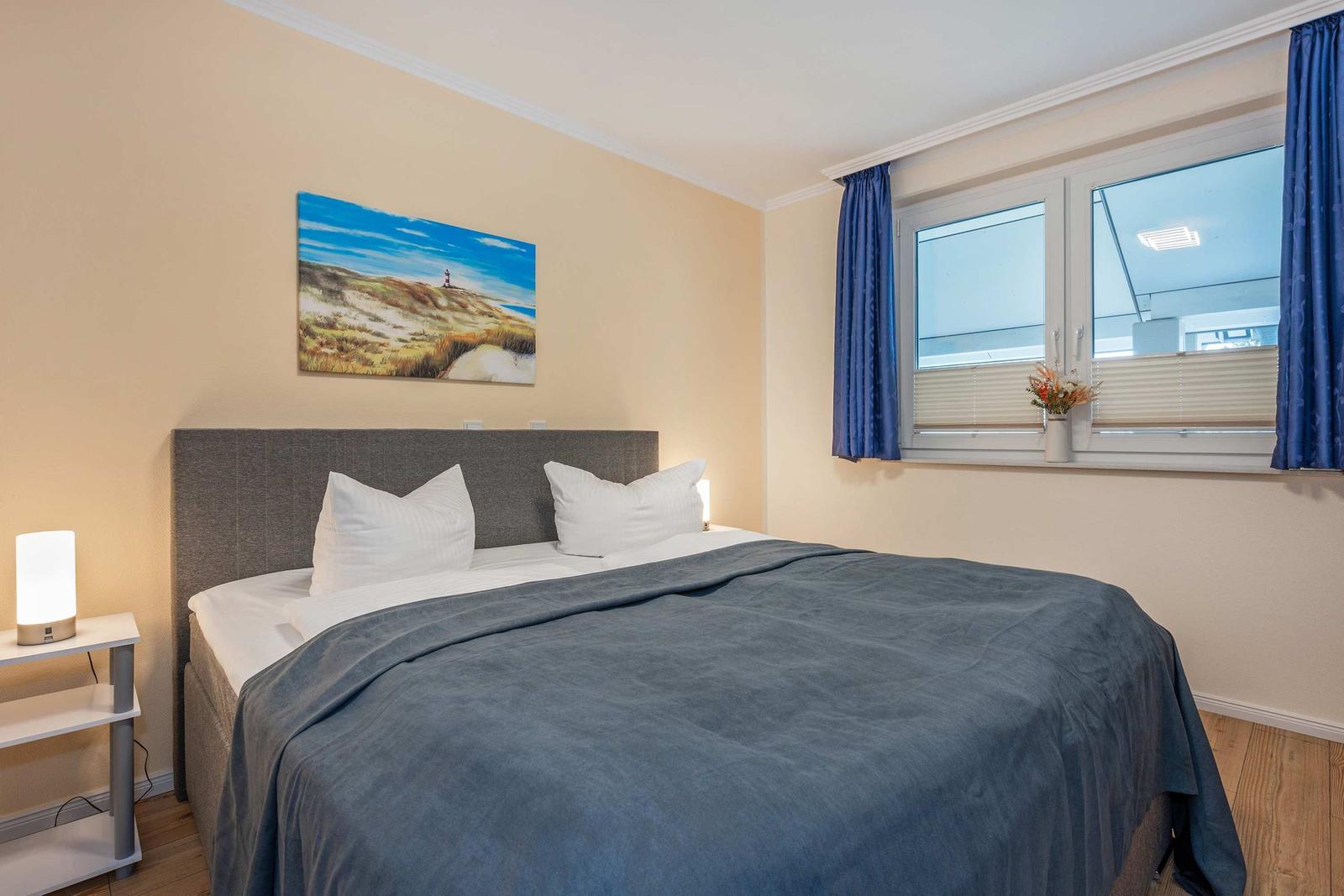 Schlafzimmer mit Bett, Wandbild und Fenster mit Vorhängen.
