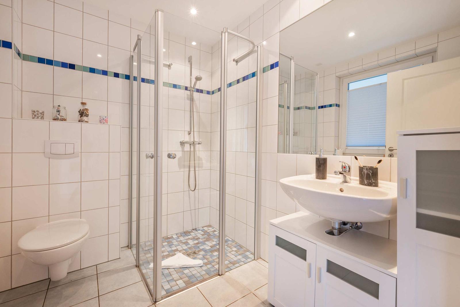 Badezimmer mit Dusche, Waschbecken und Toilette. Weiße Fliesen mit blauer Zierleiste.