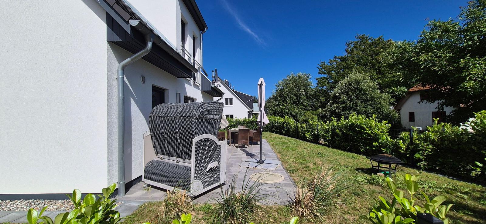 Terrasse mit Sitzgruppe, Sonnenschirm und Grillplatz im Garten. Hintergrund: Wohnhaus und Bäume.